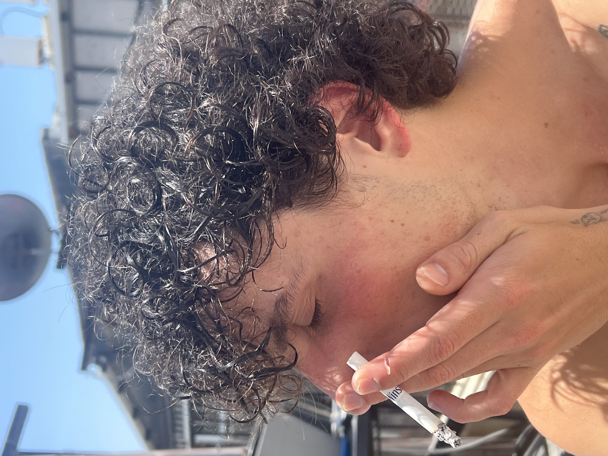 A close-up, profile shot of a young person with curly, dark hair is captured outdoors on a sunny day. The individual is holding a lit cigarette with their right hand, bringing it towards their face. Their eyes are closed, and their skin appears flushed, possibly from the sun. The background suggests an urban environment, with buildings and structures visible against a clear blue sky. The composition focuses on the person's face and hand, highlighting the act of smoking.
