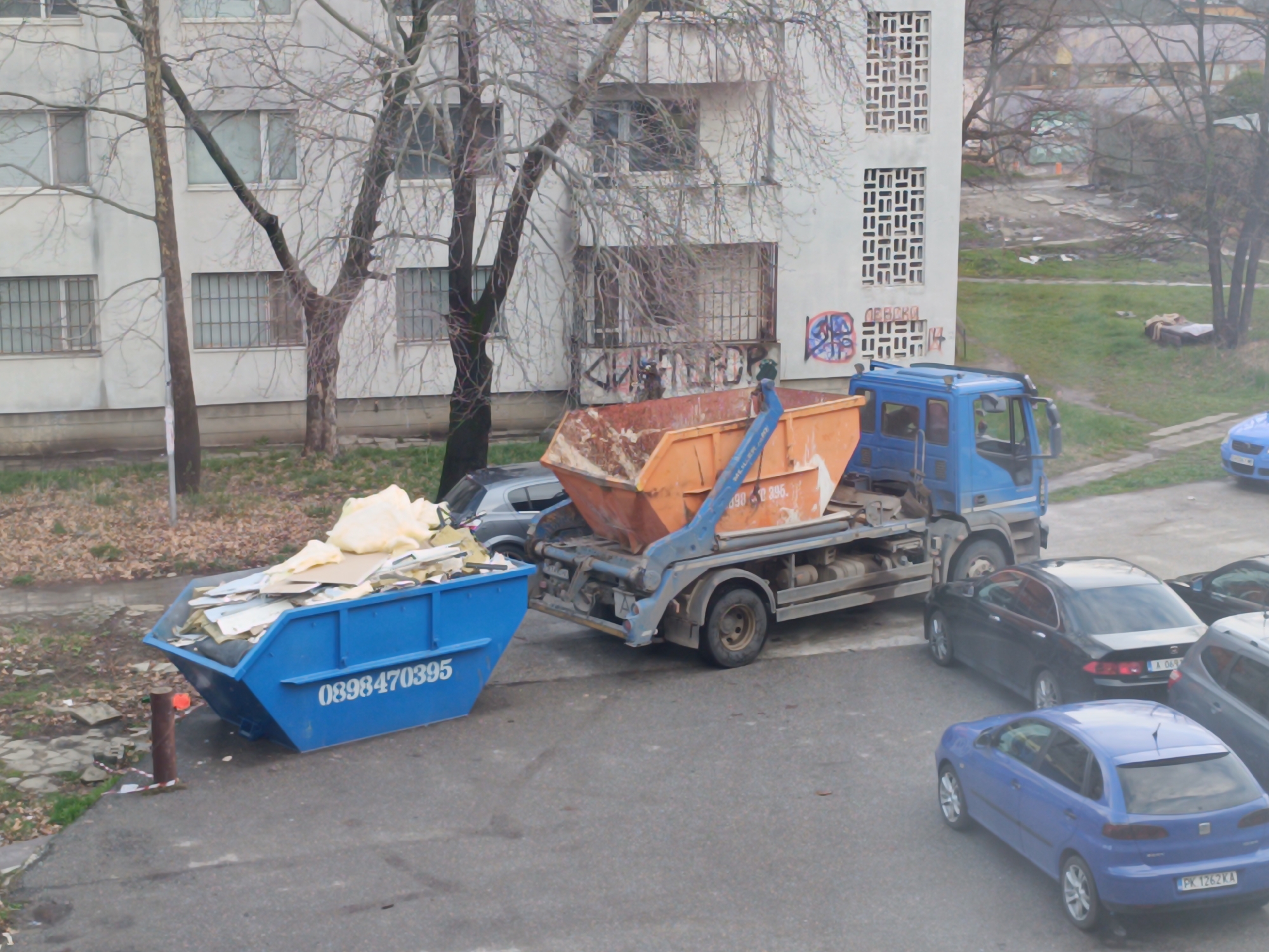 A blue skip loader truck is depicted in an urban environment in Sofia, Bulgaria, actively engaged with waste skips. The truck, positioned on an asphalt surface, has its lifting mechanism extended, holding an orange metal waste skip partially filled with construction debris, including yellow insulation material and cardboard. To the left of the truck, a large blue metal skip rests on the asphalt, overflowing with similar construction refuse. This blue skip bears the white number "0898470395" on its side.
Several passenger vehicles are parked on the asphalt. A black Honda Accord, bearing a Bulgarian Sofia license plate ("A 069..."), is visible behind the truck. In the foreground, a blue hatchback with a Bulgarian Pernik license plate ("PK 1262 KA") is parked, facing away. Partial views of other parked vehicles are also discernible.
In the background, a multi-story concrete building, consistent with residential architecture, features numerous windows, some secured with metal bars. The right section of this building displays decorative geometric concrete screens and Cyrillic graffiti, including the word "ДЕВСКО". Multiple bare, deciduous trees with intricate branch structures are scattered across the scene, particularly between the vehicles and the buildings. The ground consists of an asphalt parking area, patches of sparse grass, dirt, and scattered autumn leaves and debris, leading to a less maintained, overgrown area in the distant background. No individuals are visible in the immediate scene.
