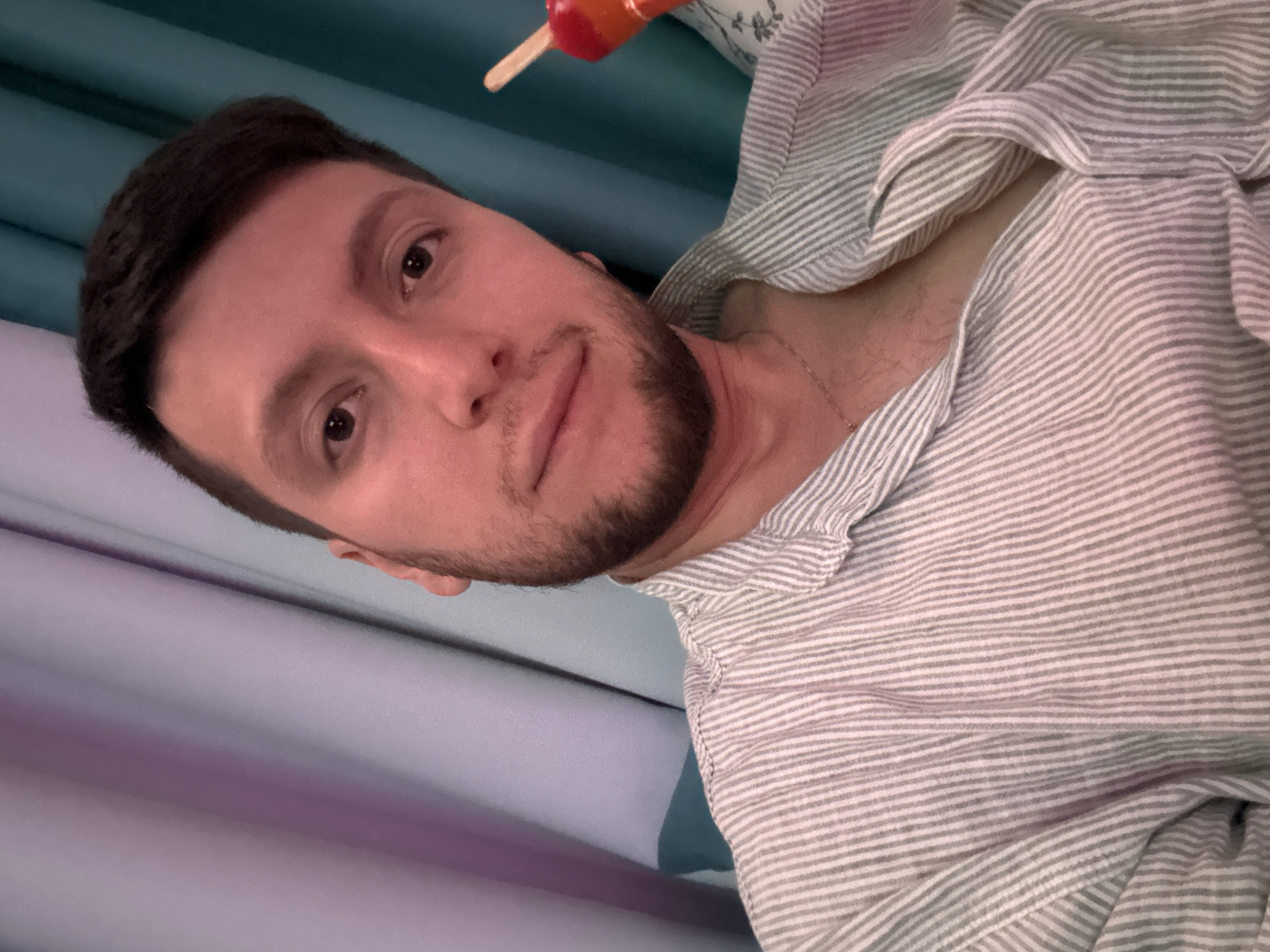 An adult male is captured in a close-up, chest-up selfie in an indoor setting in the city of Мирный, Russia. He has short dark hair, dark eyes, and a short beard or stubble. The lighting casts a warm, reddish hue on his skin. He is wearing a collared shirt with thin vertical stripes in light grey and white, unbuttoned at the top to reveal a thin metallic chain necklace and some chest hair. He looks directly at the camera with a neutral to slightly relaxed expression. In his right hand, visible on the right side of the frame, he holds a red and orange graduated popsicle or candy on a wooden stick. The background consists of draped curtains, with a light purple section on the left and a teal-blue section on the right, both exhibiting vertical folds.