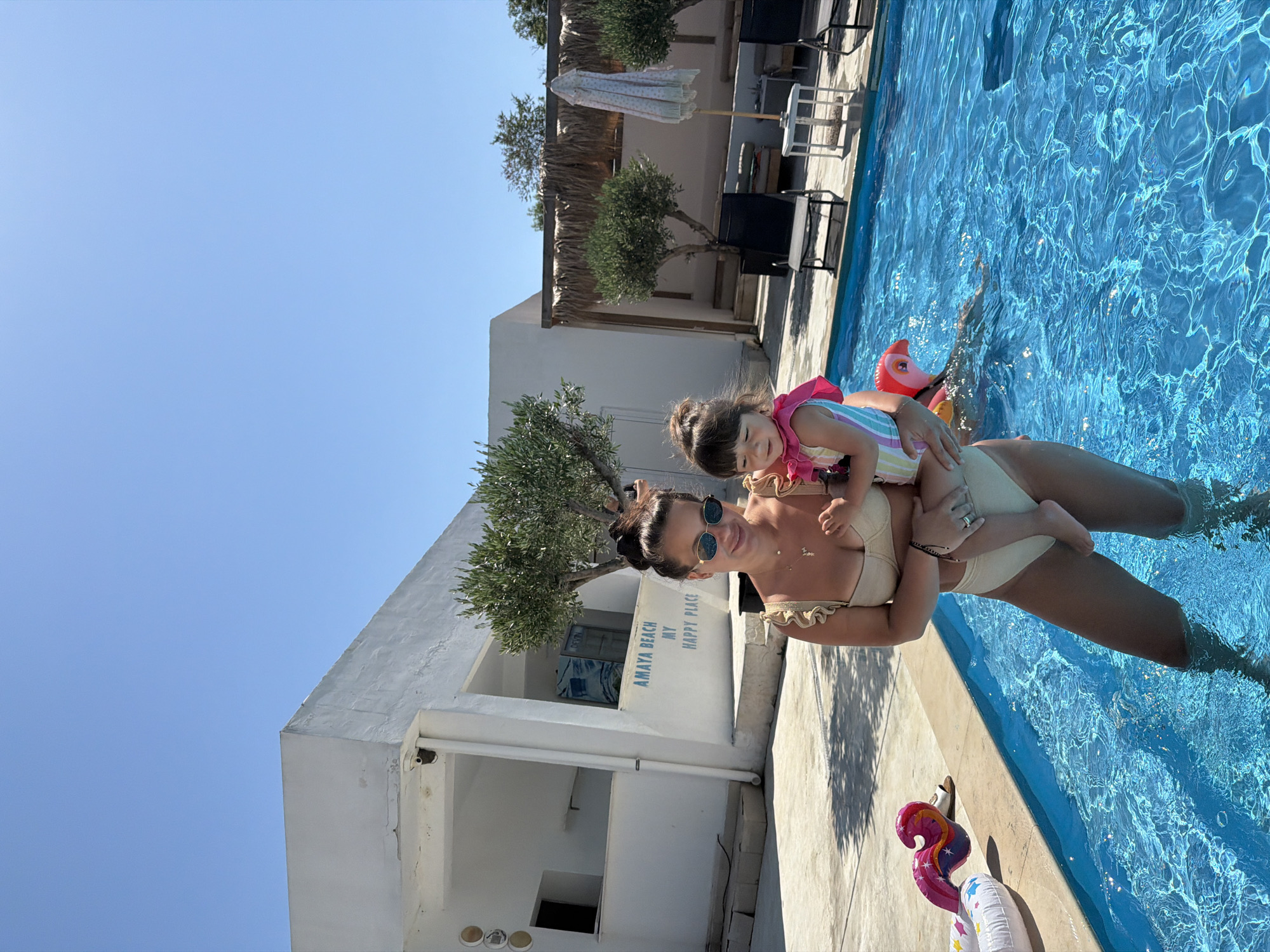 A woman and a young child are in a swimming pool. The woman is wearing a beige bikini and sunglasses. The child is wearing a striped swimsuit and a pink life jacket. The child is on the woman's shoulders, and both appear to be smiling. The pool is filled with bright blue water, and sunlight reflects off the surface. In the background, there is a white building with some olive trees and thatched roof structures. Beach chairs are visible near the pool. There is also a pink flamingo inflatable toy and a white inflatable ring with stars. The sky is clear and blue. The scene suggests a sunny day at a resort or hotel. Visible text on the building reads "AMAYA BEACH MY HAPPY PLACE". The context suggests this is in Burgas, Bulgaria.