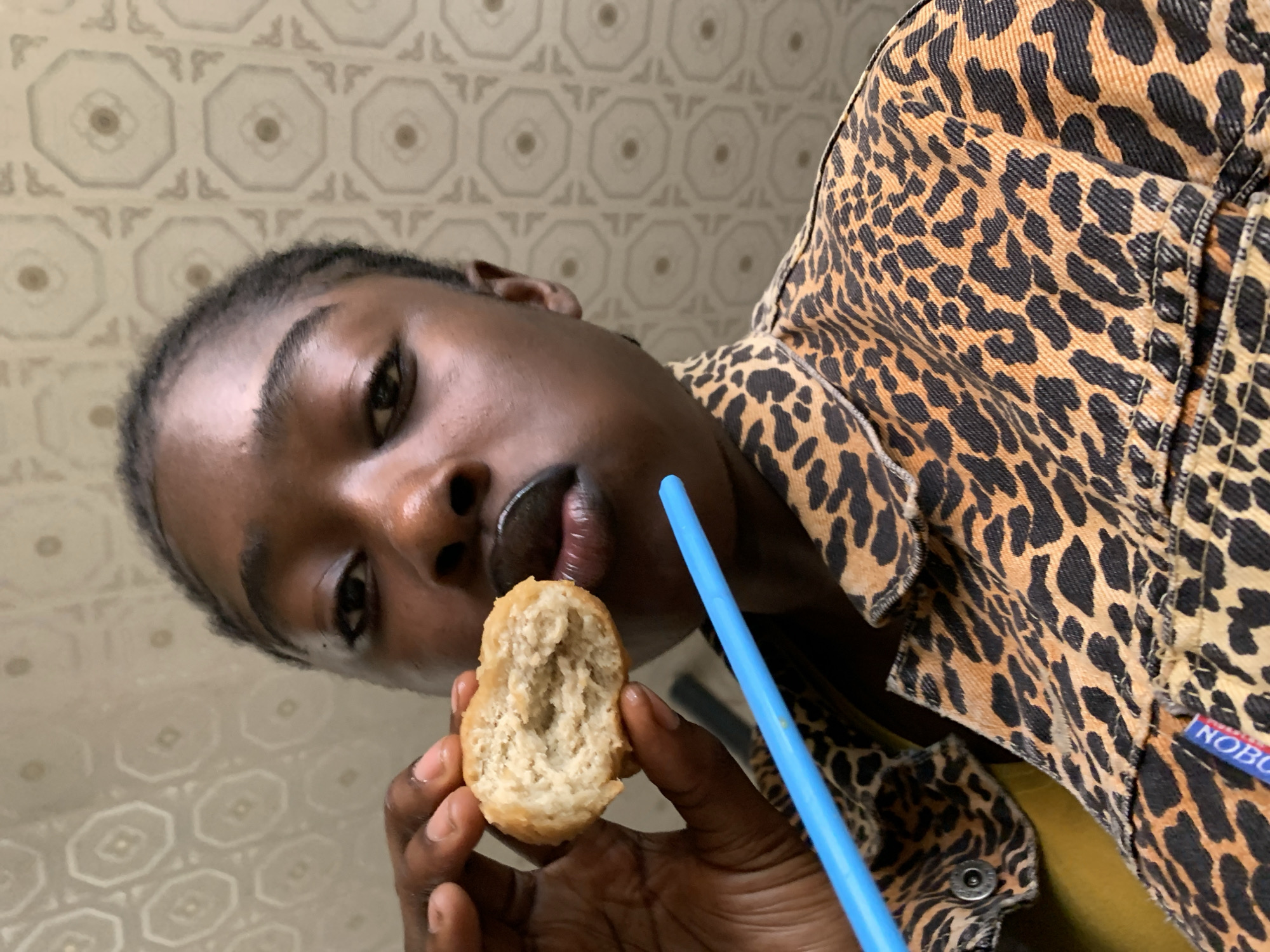 A close-up, medium shot shows a young Black person holding a bitten piece of food to their mouth. The person's face is turned slightly to the right, with dark hair pulled back and styled in braids. They are wearing dark lipstick. A bright blue straw is visible in front of their mouth, and a portion of a leopard-print jacket is visible on their body. The background features a light-colored wall with a repeating octagonal pattern. The image provides no information about the city of Kabong, Nigeria, or any specific location within it.