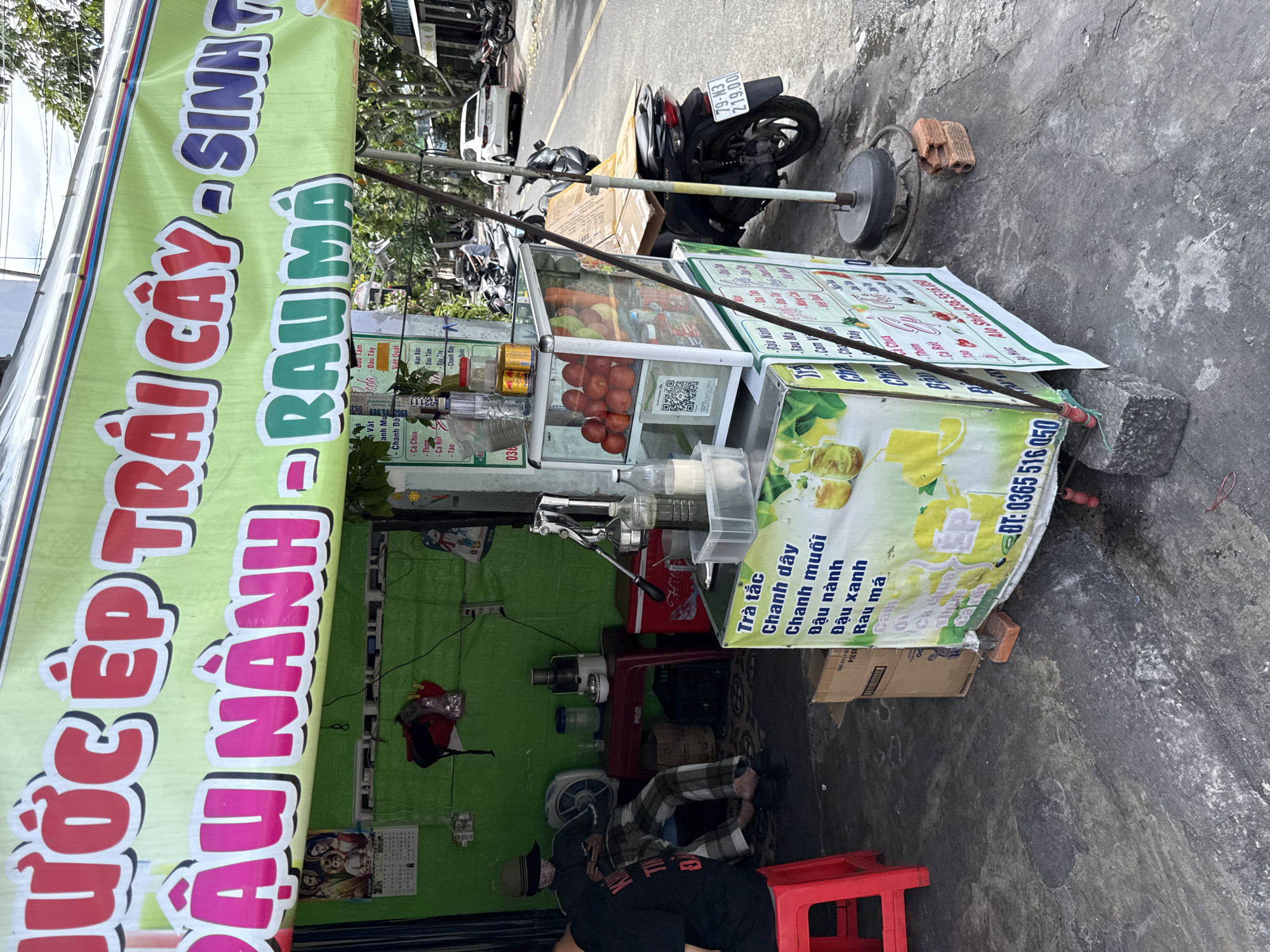 The image depicts a street-side juice stand in Nha Trang, Vietnam. A large green banner overhead reads "NƯỚC ÉP TRÁI CÂY - SINH TỐ" and "ĐẬU NÀNH - RAU MÁ," indicating it sells fruit juices, smoothies, soy milk, and pennywort juice.
In the foreground, a red plastic stool is visible. To the left, a person wearing a hat and checkered pants sits on a chair, looking at a mobile phone. Next to them is a white electric fan and a red table holding a juicer and a red cooler.
The juice stand itself is a mobile cart with a glass display case filled with red tomatoes and other produce. Various beverages are displayed on shelves. A printed menu lists drinks such as "Trà tắc," "Chanh dây," "Chanh muối," "Đậu nành," "Đậu xanh," and "Rau má." A QR code is also visible on the cart.
In the background, motorcycles are parked along the street, and a white car is driving by. The overall environment appears to be an urban street scene on a bright day.