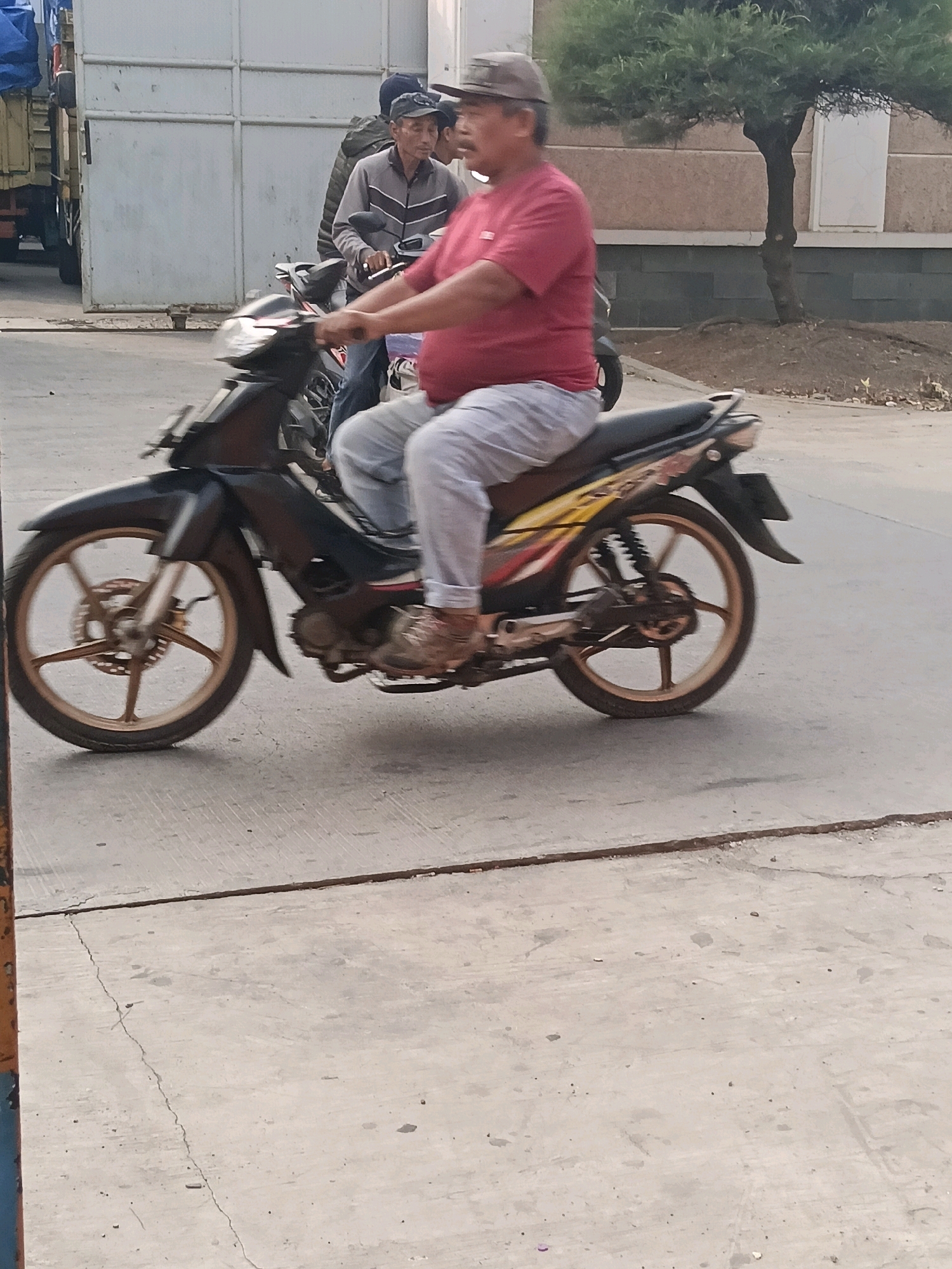 The image shows a man riding a motorcycle on a concrete road. The man is wearing a red shirt, gray pants, and a hat. He is in the process of riding the motorcycle, with his legs on the footpegs and his hands on the handlebars. The motorcycle is black with gold rims and has a license plate. In the background, there is a building and a tree. The overall environment appears to be an outdoor setting, possibly a street or parking lot in the city of Belimbing, Indonesia.