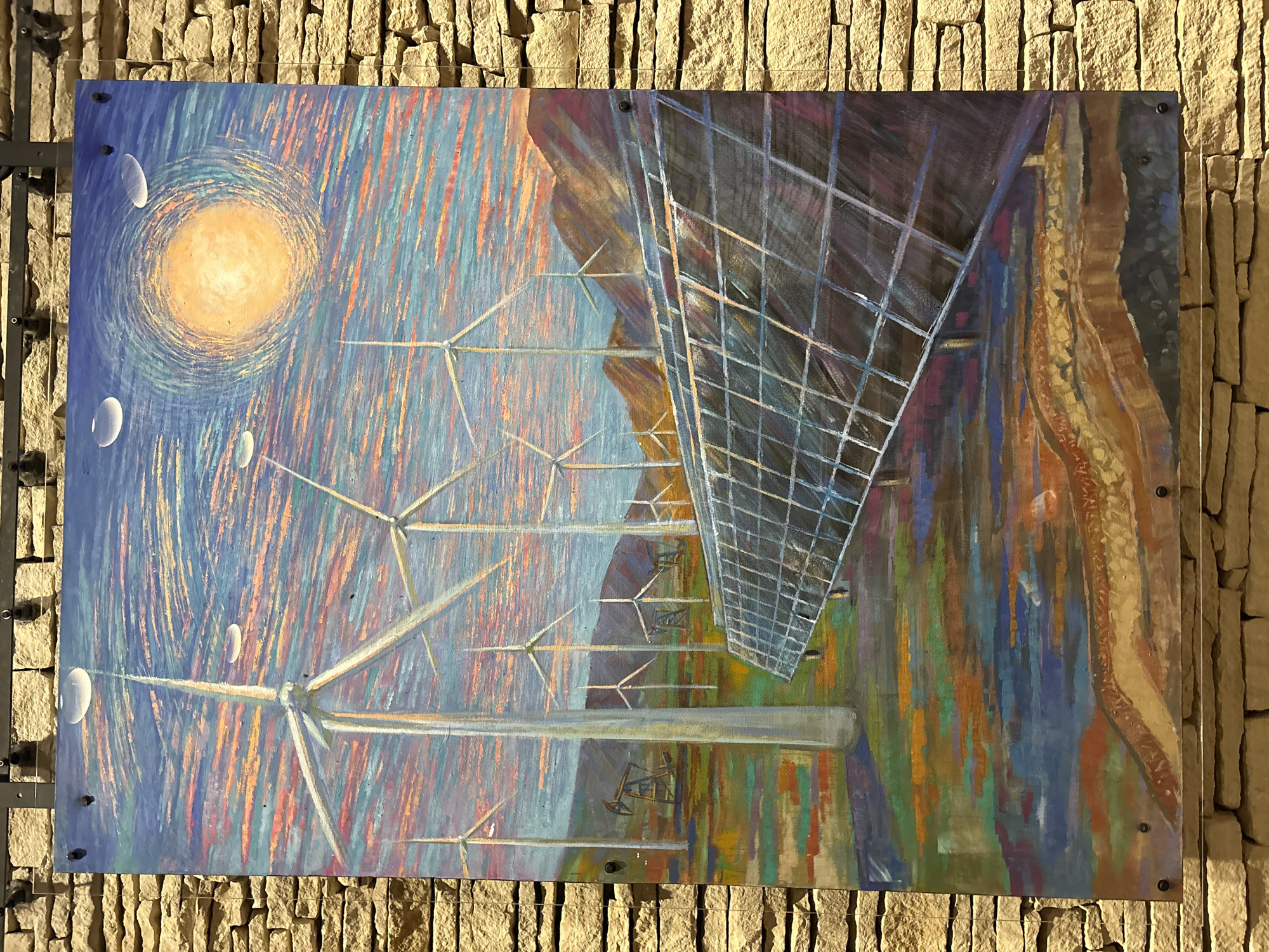 The image displays a painting mounted on a stone wall. The painting depicts a landscape with wind turbines and solar panels, suggesting a focus on renewable energy. The sky is painted with swirling strokes of orange, pink, and blue, with a bright yellow orb representing the sun. Several wind turbines are visible in the middle ground, with what appear to be oil pump jacks in the distance. In the foreground, a large array of solar panels is depicted at an angle. The overall style of the painting is impressionistic, with visible brushstrokes and vibrant colors. The location is identified as Unknown, United States of America.