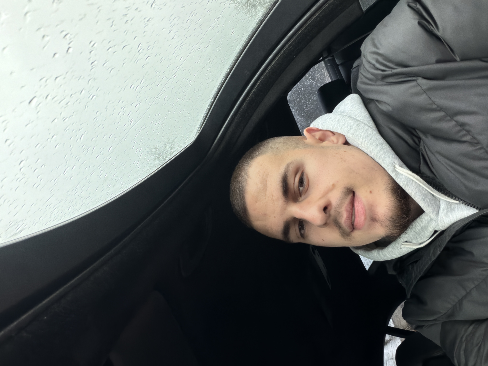 A person is photographed inside a vehicle with a sunroof on a rainy day. Raindrops are visible on the sunroof glass. The person has short hair, facial hair, and is wearing a gray hooded sweatshirt under a black puffer jacket. The vehicle's interior is dark. There is no specific information to indicate that the photo was taken in Plovdiv, Bulgaria.