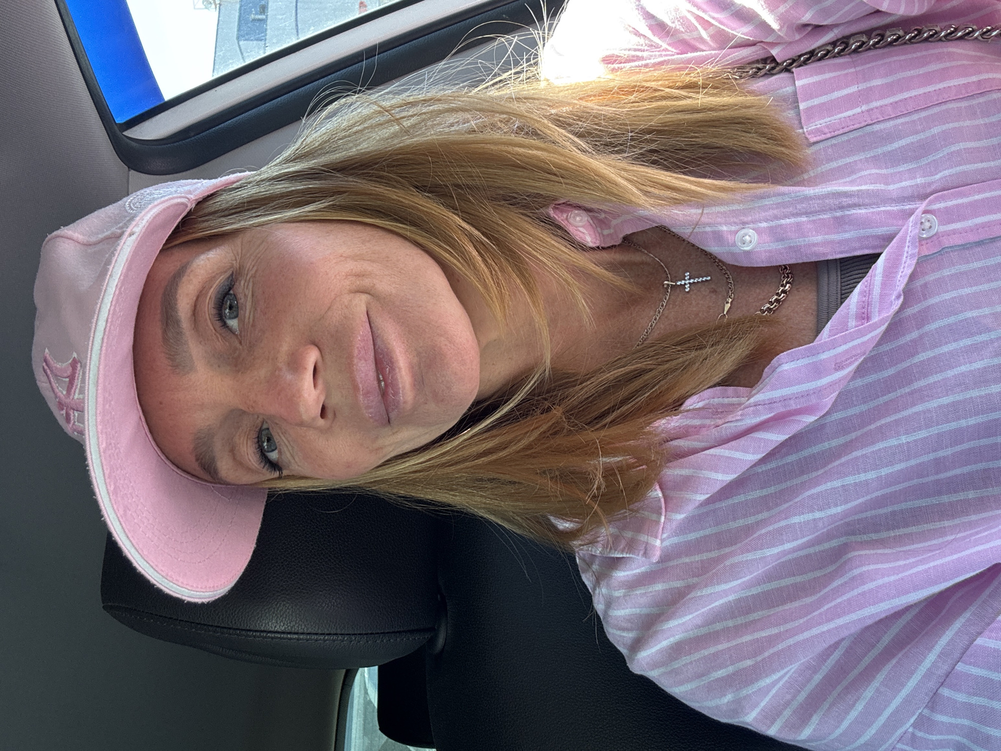 A woman is captured in a close-up shot, presumably a selfie, inside a vehicle in Saint Petersburg, Russia. Her head is tilted to the side as she looks directly at the camera with a subtle smile or smirk. She has long, light brown to blonde hair, blue eyes, and a light complexion.
She is wearing a casual outfit consisting of a pink baseball cap with white embroidery that is not clearly legible, and a pink and white striped button-up shirt worn over a grey undershirt. Around her neck, she has two silver chain necklaces, one featuring a small cross pendant. The bright, natural light entering through a window suggests it is daytime.
The setting appears to be the interior of a car or similar vehicle, indicated by a black leather headrest behind her and a window with a dark frame and blurred exterior view in the upper left background. The overall impression is one of a relaxed, candid moment.