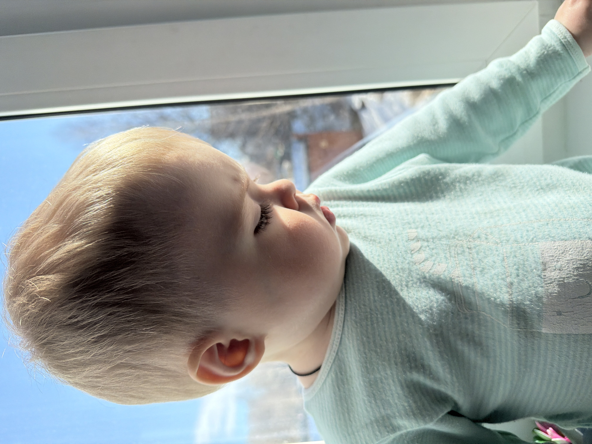 A young child, appearing to be a toddler with blonde hair and fair skin, is depicted in a close-up, three-quarter profile, looking downwards with eyes cast low or closed. The child is wearing a light mint-green, horizontally striped long-sleeved top with a white collar trim; a thin dark cord is visible around the neck. A faint light-colored graphic is present on the chest of the top. The scene takes place indoors at a window in Votkinsk, Russia. The child's right arm is visible, with the hand resting on a white windowsill. Bright natural light illuminates the child from the left. Outside the window, under a clear blue sky, bare tree branches, patches of snow or light ground cover, and a distant building with a reddish-brown roof are visible, indicating an outdoor, possibly residential, setting during daylight hours.