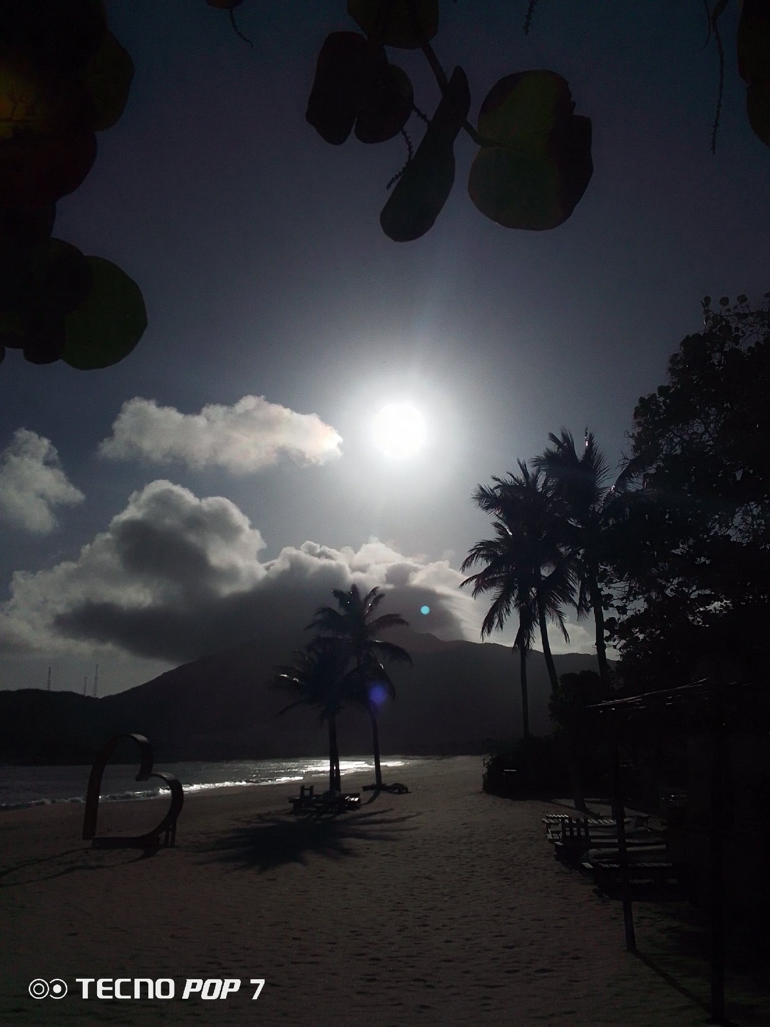 The image captures a beach scene in Pedro González, Venezuela, likely during daylight hours with a bright sun obscured by some clouds. The sky has dark areas due to the camera adjusting to the bright sun.
In the foreground, a sandy beach leads towards the water's edge, and in the midground, there are several palm trees, and a mountain in the distance. To the left, a heart-shaped structure stands on the beach. There are also what appear to be chairs or benches.
The image is somewhat dark due to the bright source of light, causing underexposure and obscuring details. Silhouetted leaves and branches are visible in the top corners. A "Tecno Pop 7" watermark is present in the bottom left corner.