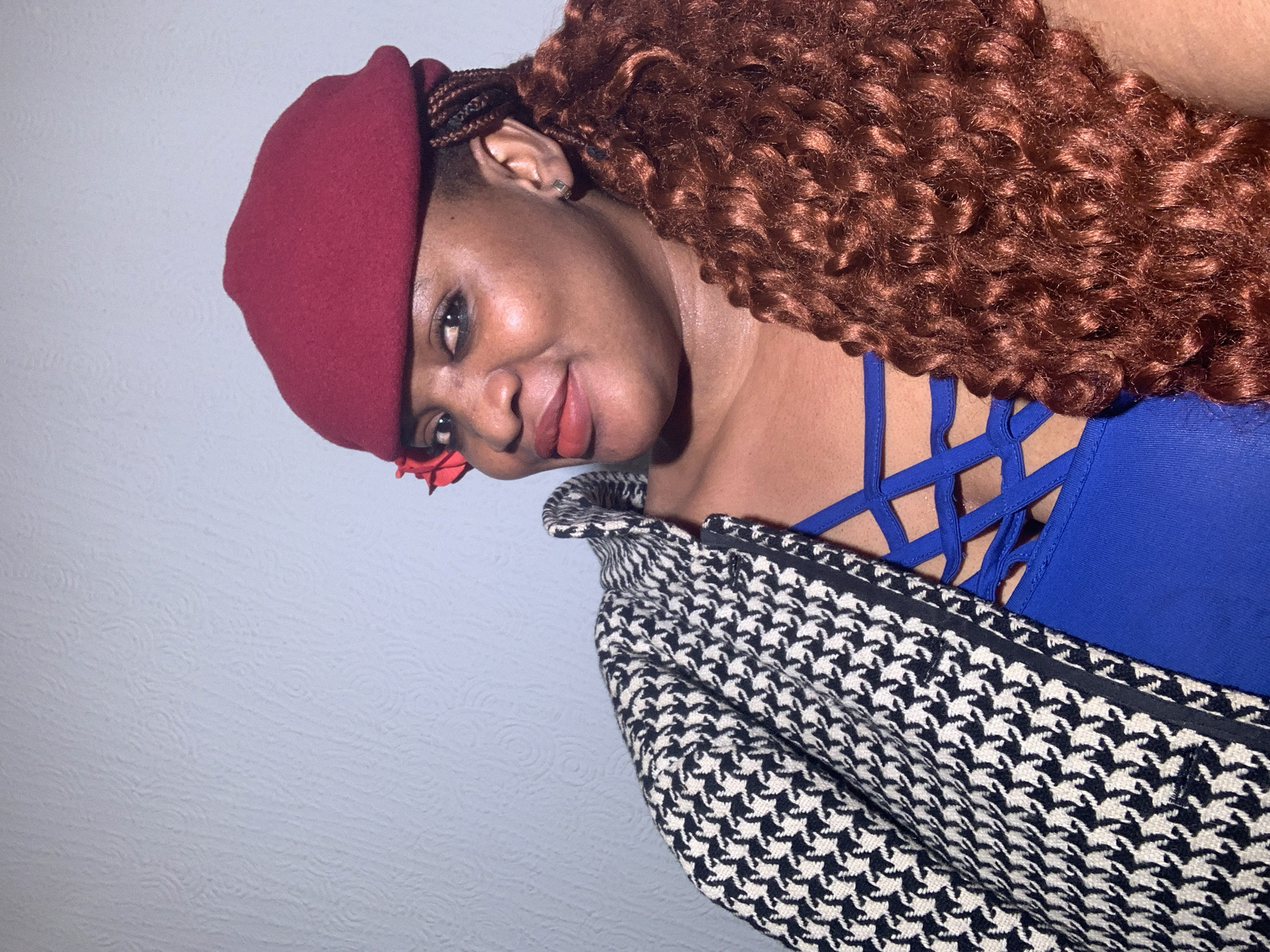 A woman is captured in a close-up portrait, tilting her head and looking directly at the viewer with a calm, pleasant expression and a slight smile. She has dark skin and is wearing a deep red beret on her head, partially covering her hair. Her hair is voluminous, curly, and a reddish-brown color, with some braided sections visible near her left ear. A small silver-toned earring adorns her ear. A small red flower-like accessory is visible near her left cheek.
She is dressed in a vibrant blue top featuring distinctive criss-cross strap detailing at the neckline. Over her top, she wears a black and white houndstooth patterned jacket or coat, which drapes over her shoulders. The background is a plain, light-colored wall with a subtle textured pattern, suggesting an indoor setting. The lighting appears warm, casting some soft shadows. The overall impression is a posed shot, possibly a selfie or portrait, taken indoors. There is no visible text in the image. The scene is set indoors, likely in Barakin Akawo, Nigeria.