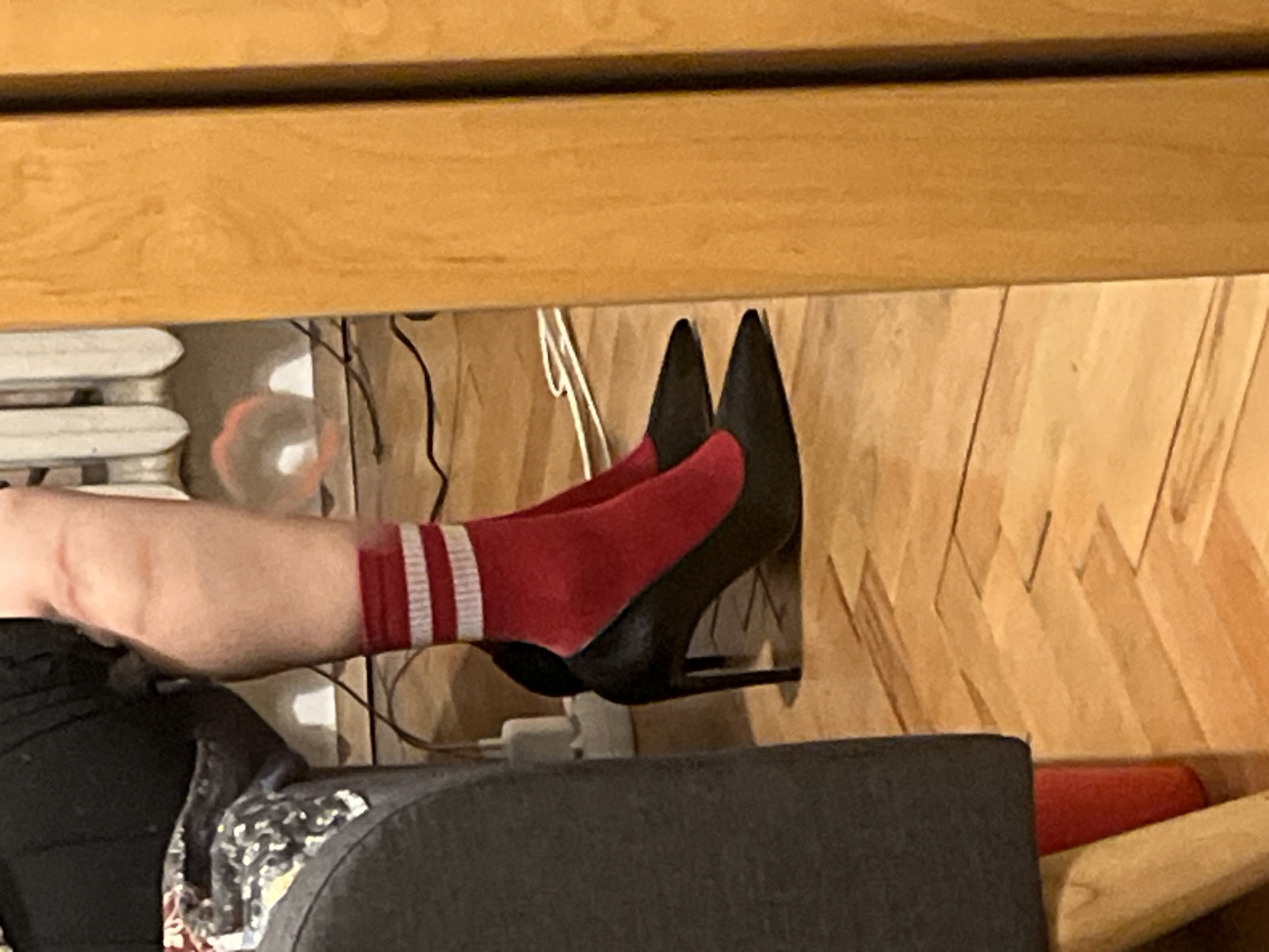 A person's leg, wearing a red sock with white stripes, extends into a black high-heeled shoe. The person is sitting on a dark gray upholstered piece of furniture. The background is a wooden wall and floor. There are cords and a white power adapter plugged in nearby. The location is not identifiable.