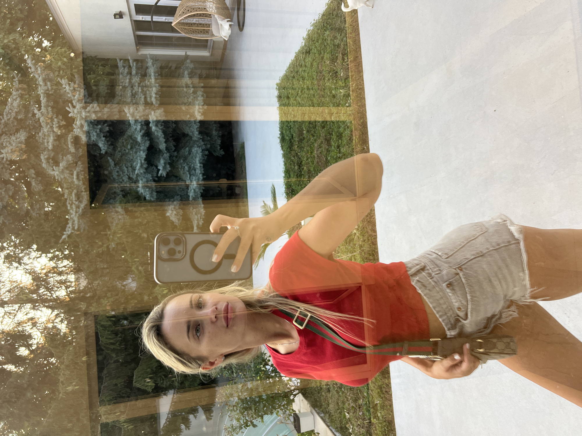 A woman with blonde hair is taking a selfie in front of a large reflective window, likely in a modern home. She is wearing a red crop top and light-colored denim shorts. A cross-body bag with a green and red strap is slung over her shoulder. The reflection in the window shows trees, the exterior of a house with a hanging chair, and a patch of green lawn. The setting appears to be outdoors, possibly a backyard or patio area. There is no indication of the specific location being the city of Fontanka, Ukraine.