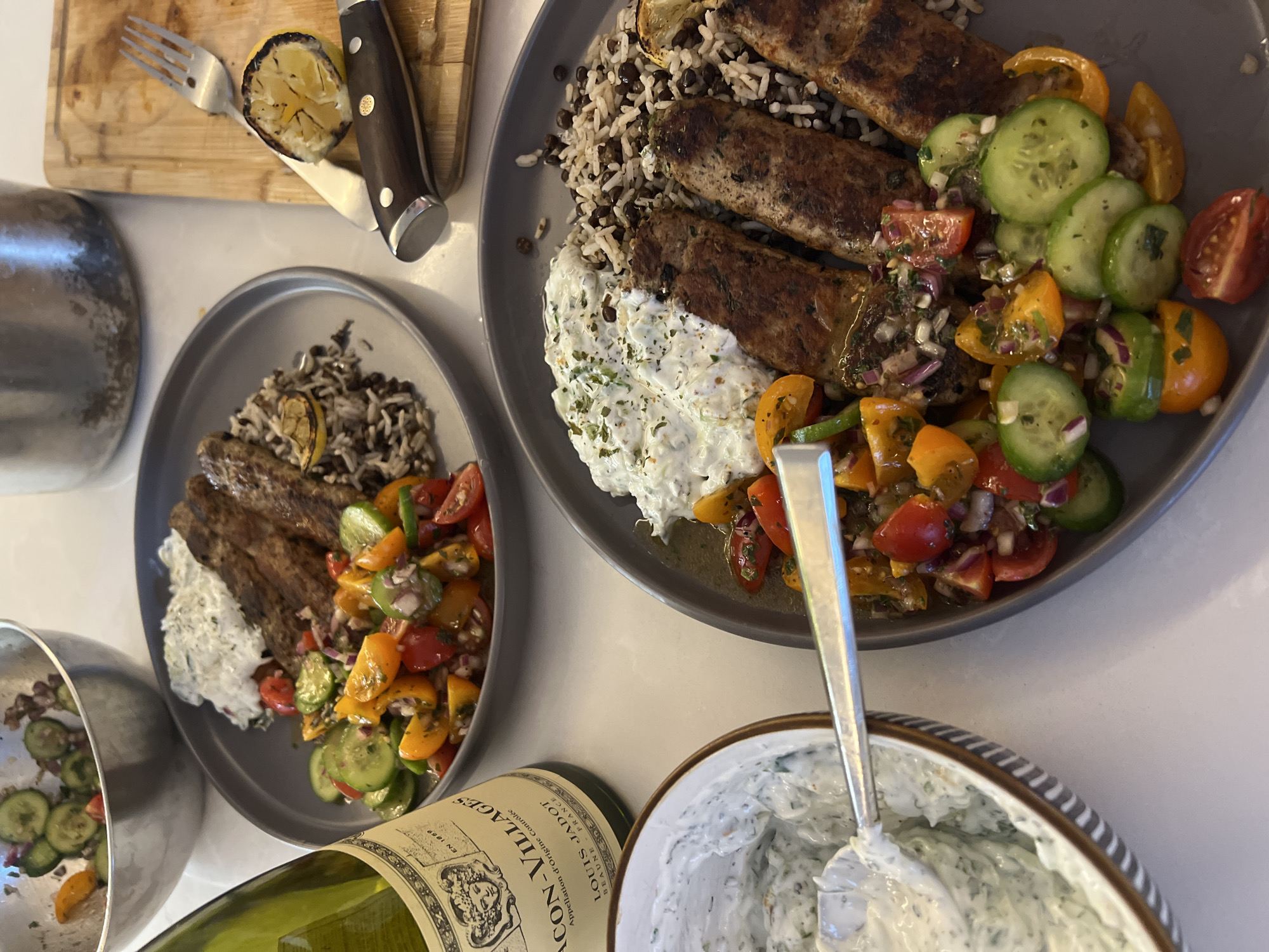 The image shows a table setting with two gray plates of food, likely in a home kitchen. The food appears to be a Mediterranean-inspired meal consisting of grilled meat (possibly lamb or chicken kebabs), rice, a cucumber and tomato salad, and a white sauce (likely tzatziki). A bowl of the white sauce with a spoon inside, and a bottle of wine are also on the table. A cutting board with a lemon wedge and a knife is in the upper left corner. The location is not visible and therefore not possible to determine it is in the city of London, United Kingdom.