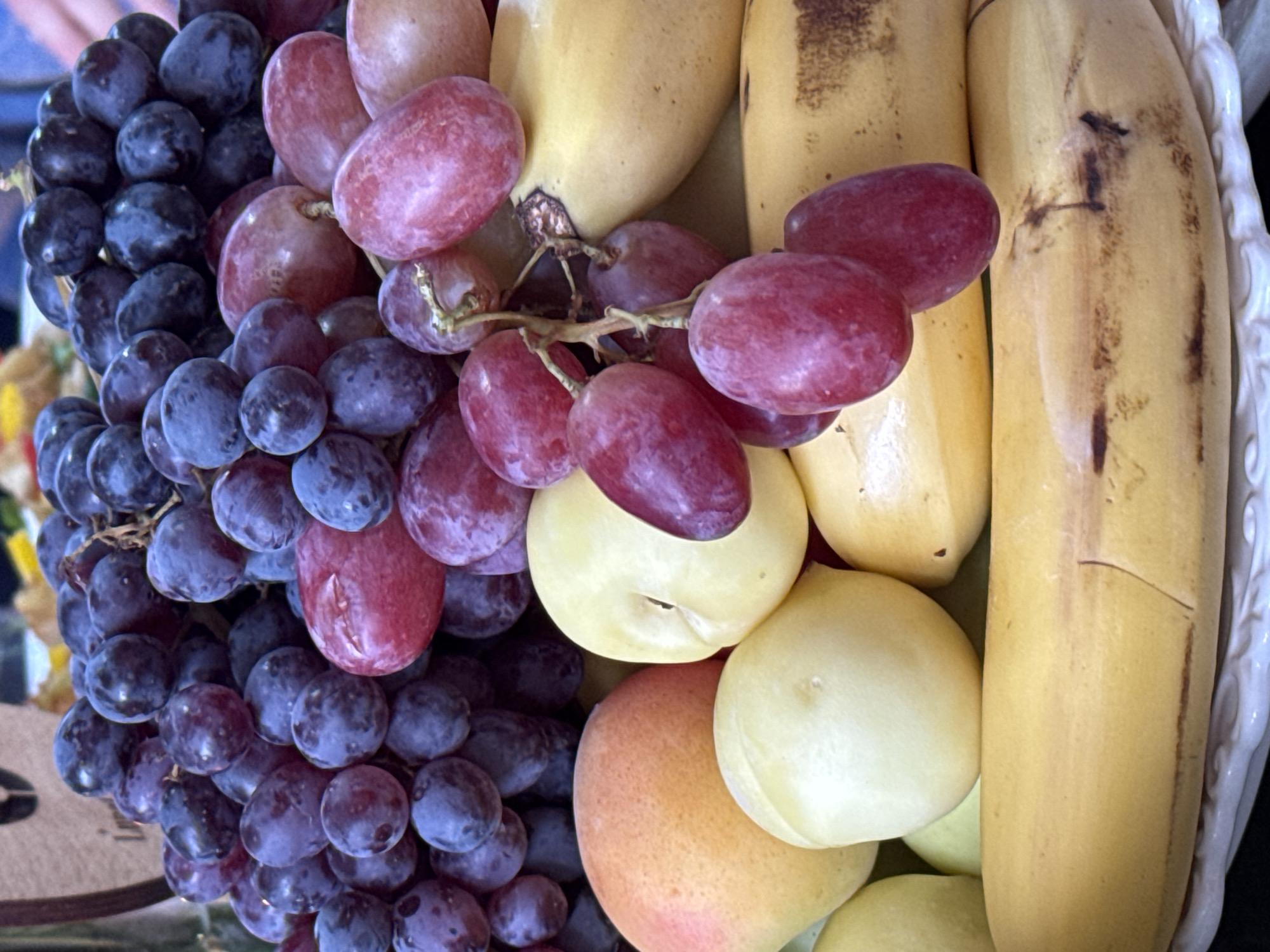 A close-up, eye-level shot shows a fruit platter filled with an assortment of ripe fruits. Dominating the composition are clusters of deep purple and red grapes, their round forms packed tightly together. Interspersed among the grapes are smooth-skinned, pale yellow fruits, likely plums or apricots, with visible indentations. Toward the bottom of the frame, several long, yellow bananas with scattered brown spots are visible, some appearing slightly bruised. The fruits are arranged on a white, textured platter, with a dark background suggesting an indoor setting, possibly a market or a home. The lighting is bright, highlighting the vibrant colors and textures of the fruit. There is no indication of the city of Arashan, Kyrgyzstan in this image.