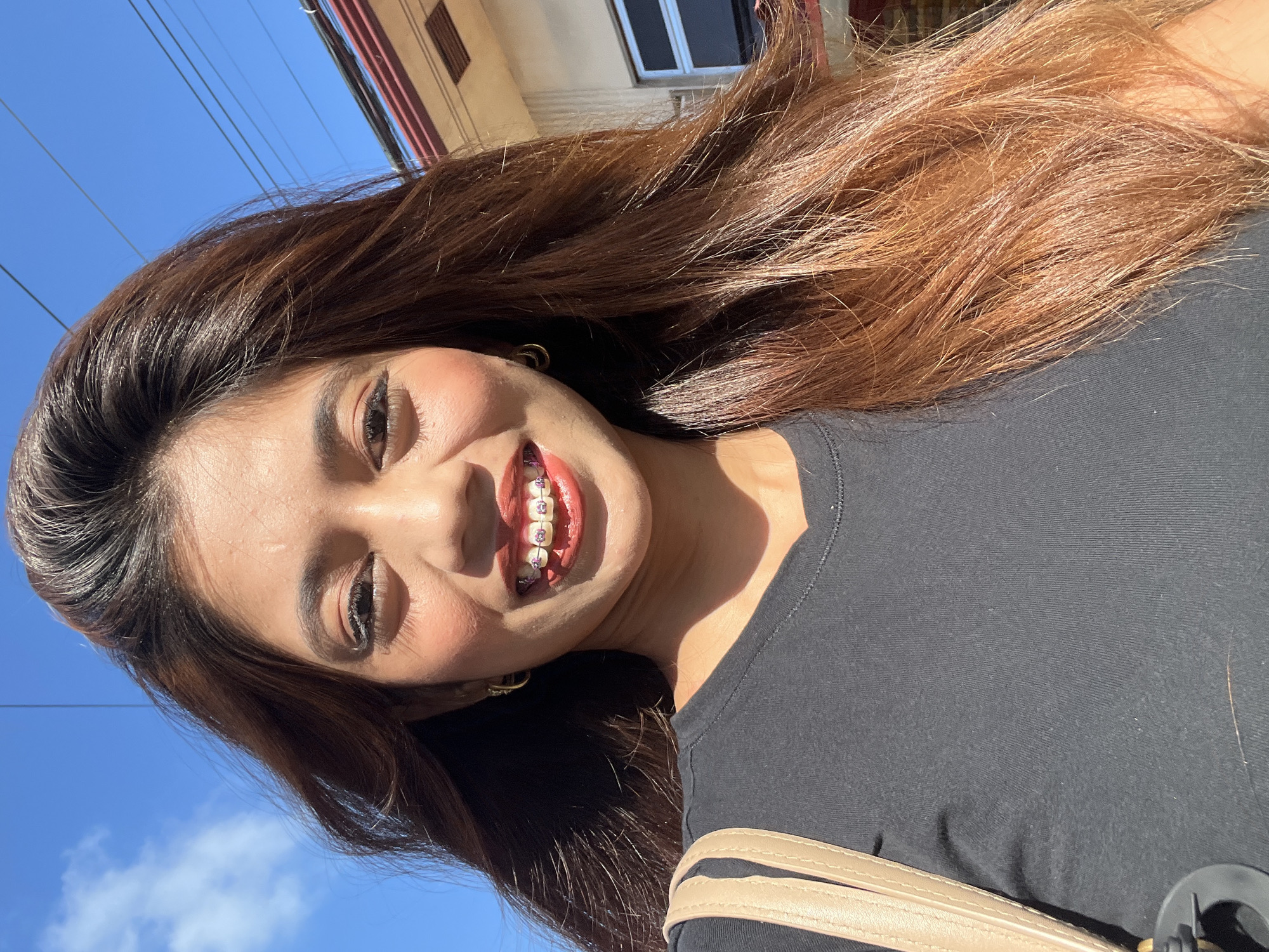 The image is a selfie of a young woman with long, reddish-brown hair and braces. She is wearing a black t-shirt and a light brown bag strap is visible over her shoulder. The background shows a clear blue sky with some clouds, power lines, and a building with red trim. The woman is smiling, showing her teeth with pink and silver colored braces. The lighting suggests it is daytime, possibly outdoors. The location is identified as Davao City, Philippines.