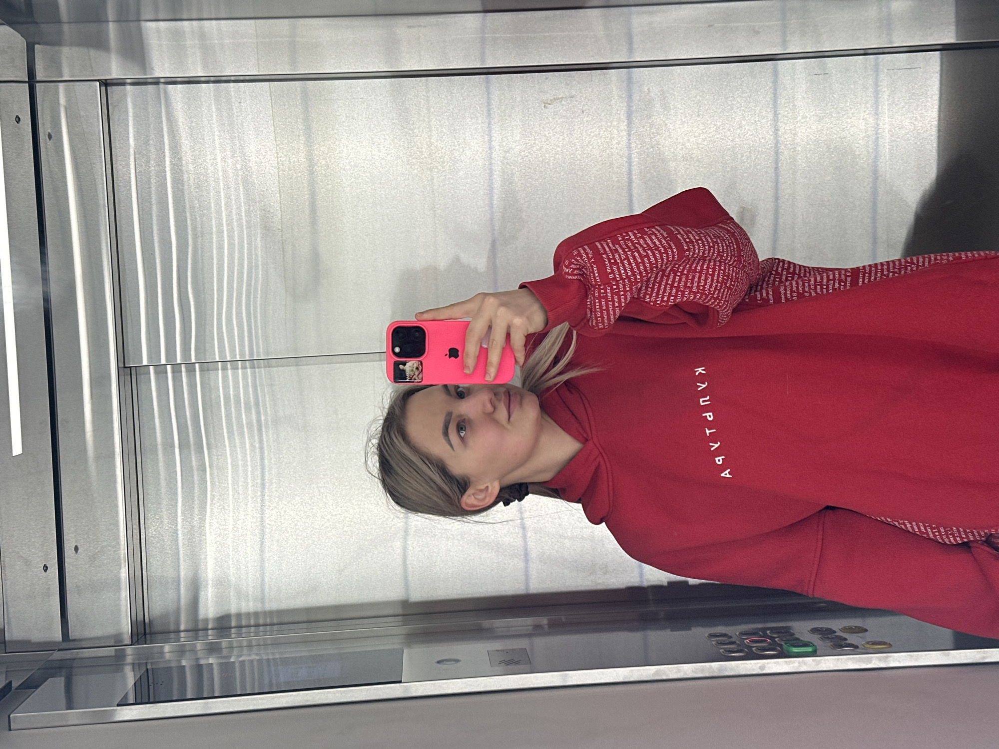 A woman is taking a selfie in an elevator. She's wearing a red hoodie with white text on the sleeves and front, and holding a pink iPhone. The elevator has stainless steel walls and a control panel with buttons. The location is unknown, and there is no mention of Budva, Montenegro in the image.