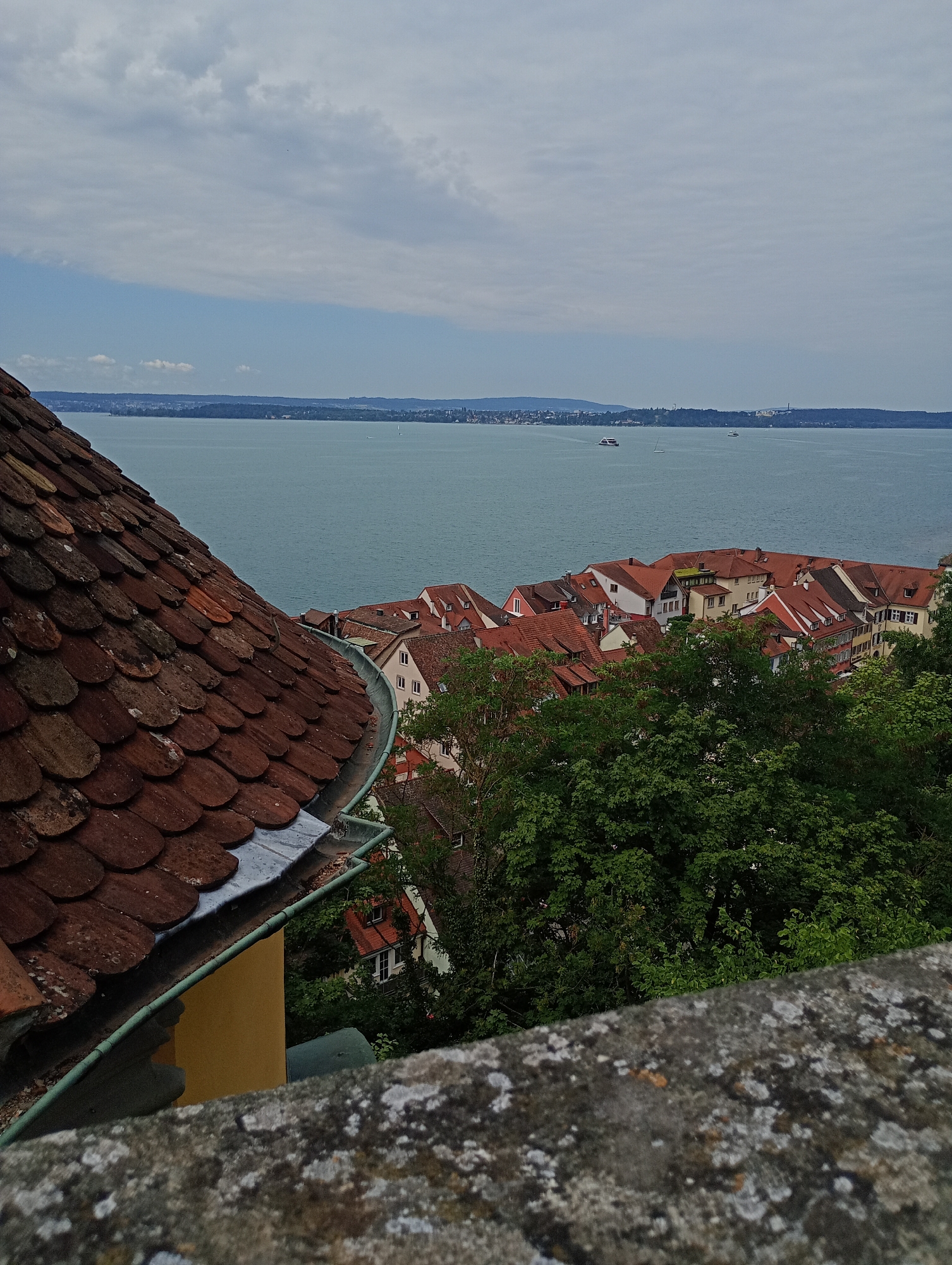 The image captures an elevated view of Meersburg, Germany, overlooking Lake Constance. In the foreground, a weathered, reddish-brown tiled roof slopes down, partially obscuring a yellow building and a grey, lichen-covered stone wall. Beyond this, a dense cluster of buildings with red-tiled roofs forms the town. The buildings are closely packed, with varying architectural styles, suggesting an old European town. Green trees intersperse among the buildings and line the foreground wall. The background is dominated by the expansive, calm blue waters of Lake Constance, with a distant shoreline visible under a cloudy sky. Several small boats, including a ferry and sailboats, are scattered across the lake. The overall impression is a picturesque, historic European lakeside town scene.