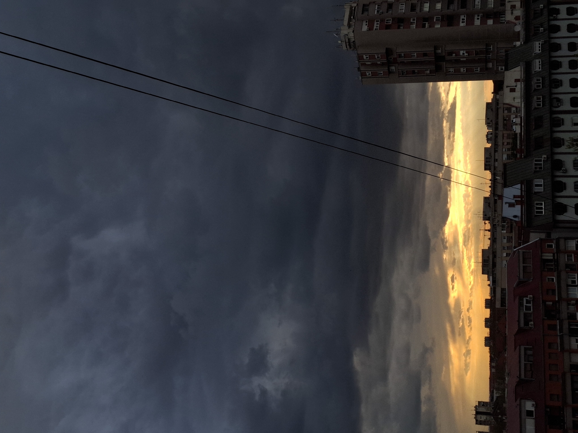 The image presents an urban landscape in Novi Sad, Serbia, captured during a dramatic sky event, likely a sunset or sunrise. The scene is dominated by a sharply contrasted sky and a dense arrangement of city buildings.
The upper and left portions of the frame are filled with heavy, dark grey and slate-blue clouds, suggesting an overcast or stormy weather condition. These dark clouds transition abruptly to a bright, intensely golden-yellow light band along the right side of the horizon, indicating the sun's low position. Two thin, dark power lines stretch diagonally across the dark cloud portion of the sky, adding a human infrastructure element to the natural phenomenon.
Along the right and bottom edges of the image, numerous multi-story buildings form a dense urban silhouette against the bright sky. These include various apartment blocks and commercial structures, characterized by multiple windows and diverse architectural styles. A tall, light-colored building with many small rectangular windows is prominent on the upper right. Further down, buildings with red rooftops and visible air conditioning units contribute to the cityscape, typical of Eastern European urban environments.
No people or specific activities are visible, emphasizing the static beauty of the moment. The photograph captures the dramatic interplay of light and shadow, highlighting the juxtaposition of turbulent skies with the enduring city structures. The overall mood is one of dramatic contrast and atmospheric intensity, showcasing the urban environment under a striking natural light display. No visible text is present in the image.