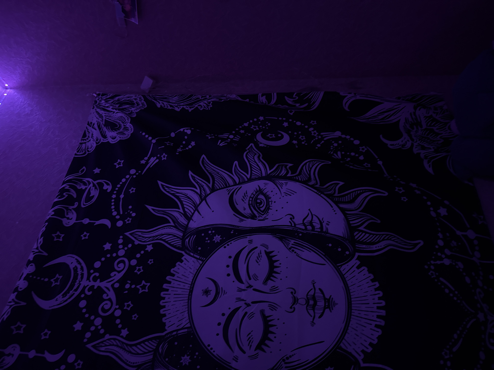 The image displays a close-up of a black tapestry with a white celestial design, illuminated by purple LED lights. The tapestry features a stylized depiction of the sun and moon, each with human-like faces. The sun is on the right, with its eye open and beams of light radiating outwards. The moon is on the left, with its eyes closed and a crescent moon symbol above its forehead. The tapestry is partially draped over a wall in a corner, where the purple lights are visible at the top, casting a strong violet hue over the scene. The walls appear textured and are also illuminated by the purple lights. The overall impression is that of a dimly lit room with a decorative tapestry as the central focus. There are no people or discernible actions visible in the image. The location in Pudomyagi, Russia is not visually identifiable from the provided image.