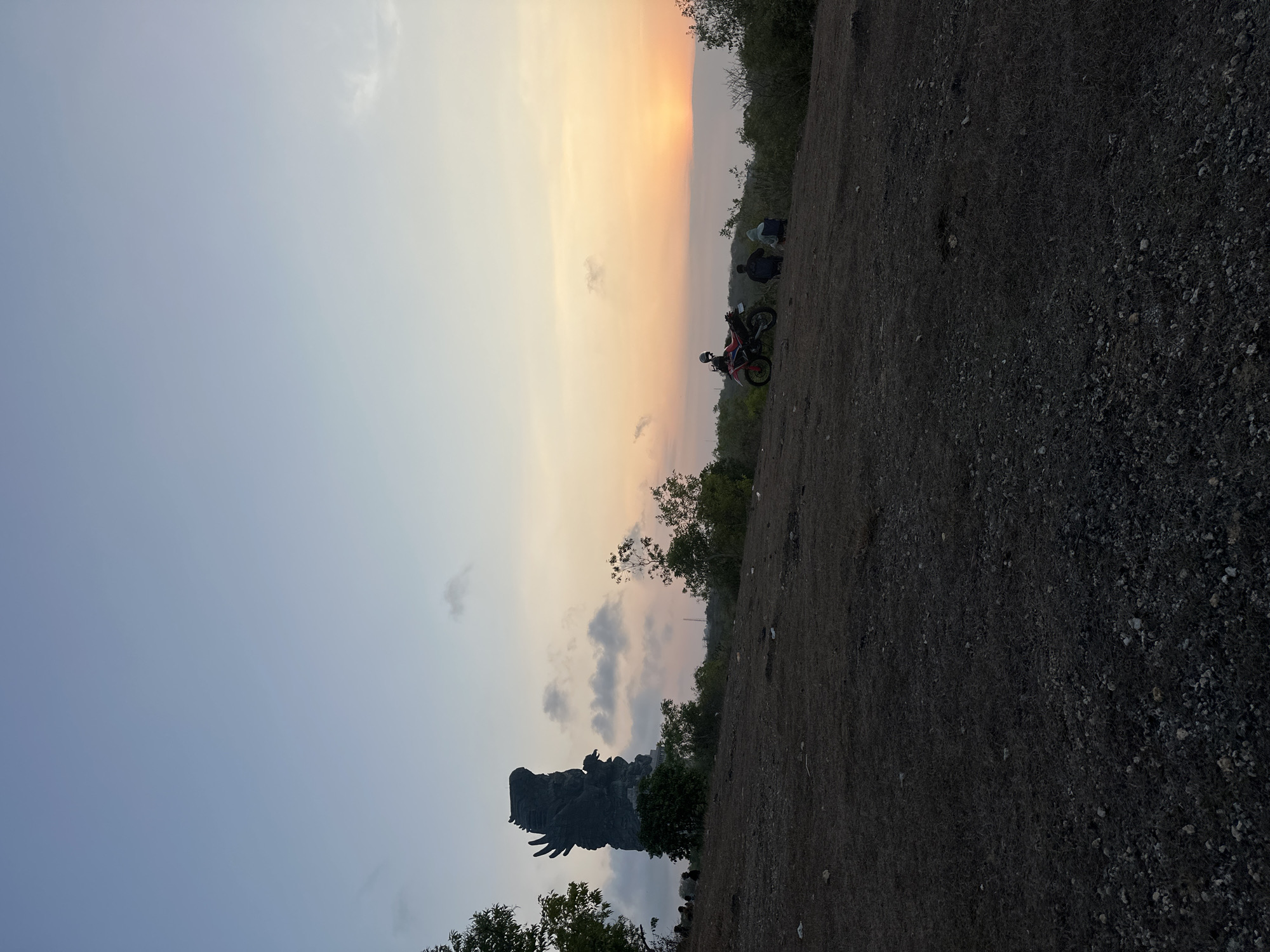 The image captures a sunset view from a hilltop in Kuta Selatan, Indonesia, likely near the Garuda Wisnu Kencana Cultural Park. In the background, a colossal statue, presumably the Garuda Wisnu Kencana statue, stands silhouetted against the sky. The sky transitions from a pale blue at the top to hues of orange and yellow near the horizon, indicating the sun is setting. Scattered clouds add texture to the sky.
In the foreground, a barren, grassy hillside slopes downwards. Several small trees and bushes are dotted across the landscape. On the right side of the frame, a red and black motorcycle is parked. Two individuals are seated on the ground near the motorcycle, facing the sunset. Another small group of figures can be seen further to the left, also on the hillside, some appearing to be walking or standing. The ground is covered in dry grass and small stones. The overall atmosphere is serene, with the natural landscape and the majestic statue creating a striking scene.