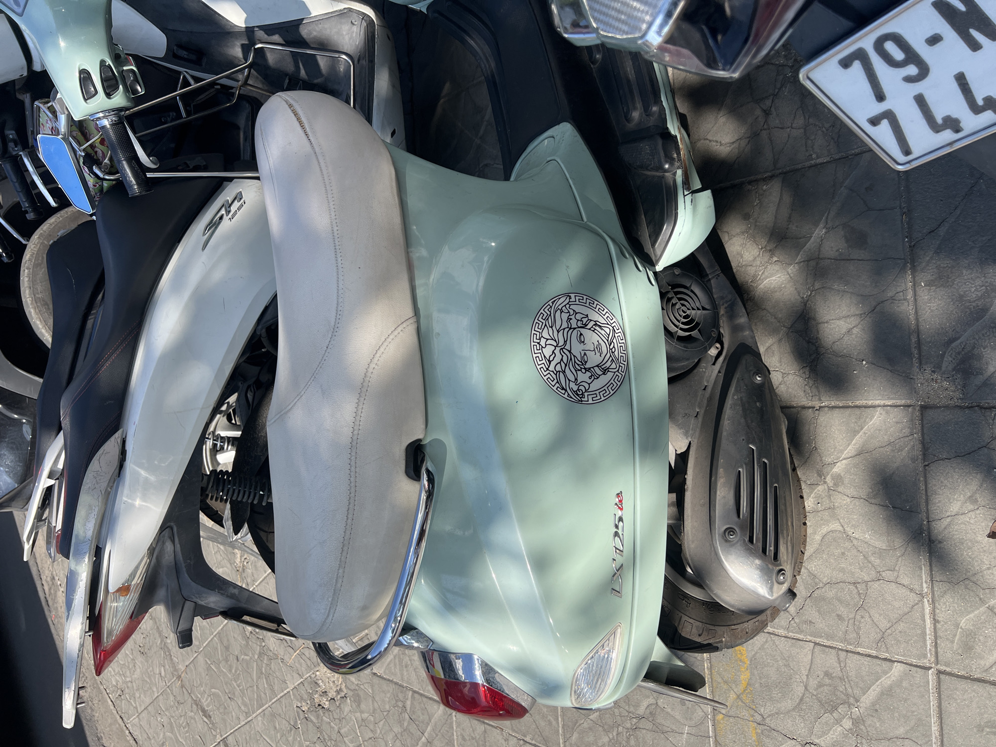 An overhead shot shows two parked scooters on a tiled pavement. The scooter on the right is light-green, with a "Versace" sticker on the front. Text "LX 125 i.e." is on the side. A small white seat is directly to the left of the light-green scooter. Part of a license plate, “79-N1 744”, is visible above and right of the front of the light-green scooter. The other scooter is silver, black, and white. The side of the fairing reads "SH 125i". The scooters appear to be parked on a street in Nha Trang Ward, Vietnam.