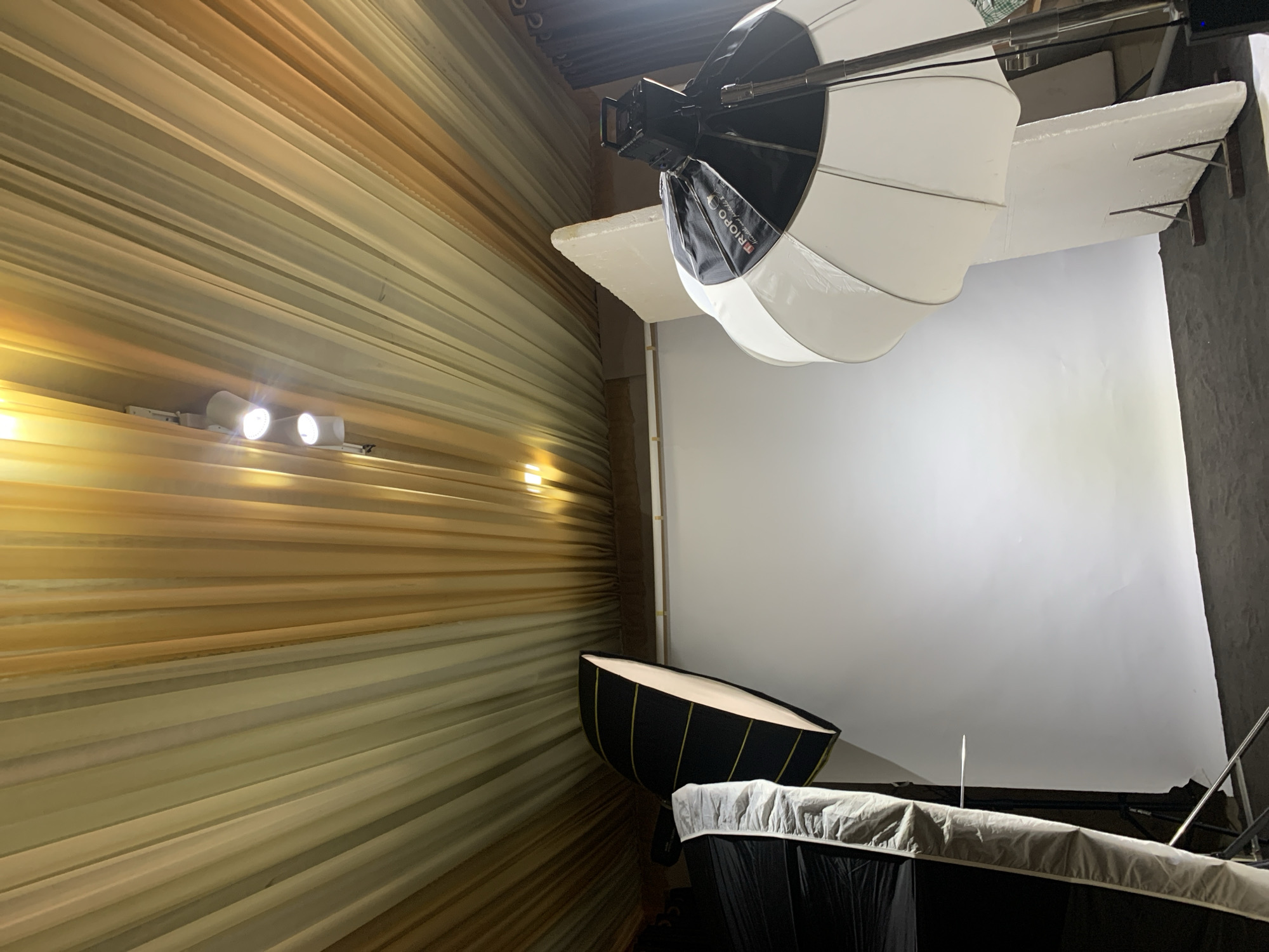 The media file shows an indoor studio setup, likely for photography or videography. A large white softbox light, with the brand "TRIPOPO" visible on a black fabric panel, is suspended from the ceiling, angled downwards. To the left, a wall is draped with flowing, pleated fabric in shades of beige and gold, illuminated by two spotlights attached to a track. In the foreground, another large, black-and-yellow striped light modifier is positioned on the floor, partially obscured by a black fabric structure. A white backdrop hangs on the right side of the scene. The overall impression is a controlled lighting environment. No people are visible. The setting appears to be a professional studio, possibly within a building in Barakin Sabawa, Nigeria. The lighting suggests artificial light is the primary illumination.