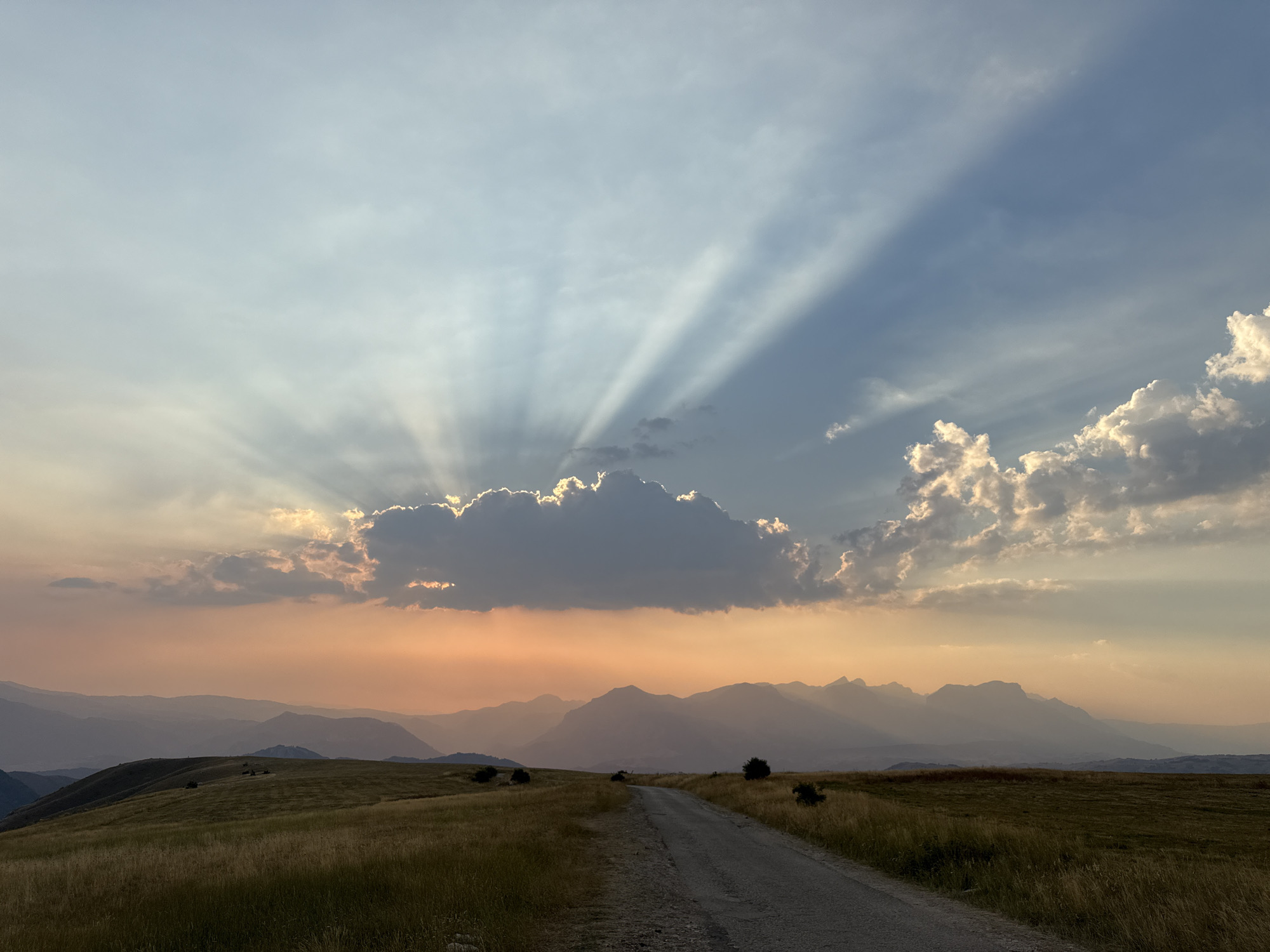 The image depicts a vast landscape at dusk, possibly in the vicinity of Boričje, Montenegro. A dirt road stretches into the distance, flanked by rolling hills covered in dry grass. In the background, a mountain range is silhouetted against a sky painted with hues of orange and blue. Dramatic sunbeams, also known as crepuscular rays, emanate from behind a prominent cloud formation, casting ethereal light across the sky. The scene is devoid of any human presence or activity. The environment appears dry and sparsely vegetated, with a few scattered bushes dotting the landscape. The overall impression is one of natural beauty and tranquility at the close of day.