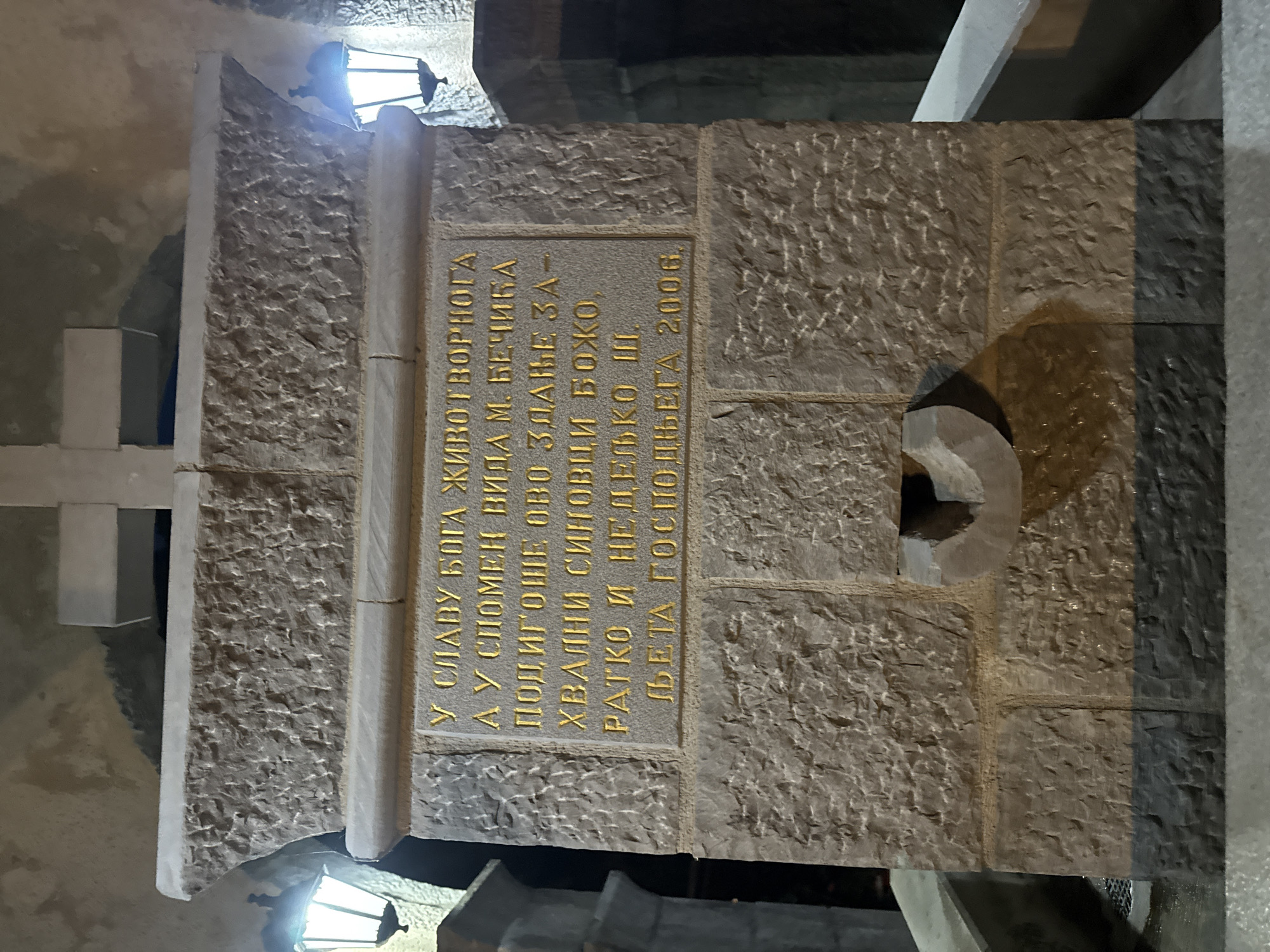 The image shows a stone monument with an inscription in gold Cyrillic text. The monument is located in Boreti, Montenegro. It is nighttime, and the monument is illuminated by two lamps on either side. The inscription reads: "У СЛАВУ БОГА ЖИВОТВОРНОГА, А У СПОМЕН ВИДА М. БЕЧИГА ПОДИГОШЕ ОВО ЗДАЊЕ ЗА- ХВАЛНИ СИНОВЦИ БОЖО, РАТКО И НЕДЕЉКО III. ЉЕТА ГОСПОДЊЕГА 2006." This translates to: "In the glory of the life-giving God, and in memory of Vida M. Bechiga, this building was erected by the grateful nephews Bozo, Ratko and Nedeljko III. In the year of our Lord 2006." The monument has a carved basin at the bottom. The overall scene suggests a memorial or a dedication, likely in a public space given the surrounding architecture which appears to be stonework and pathways.