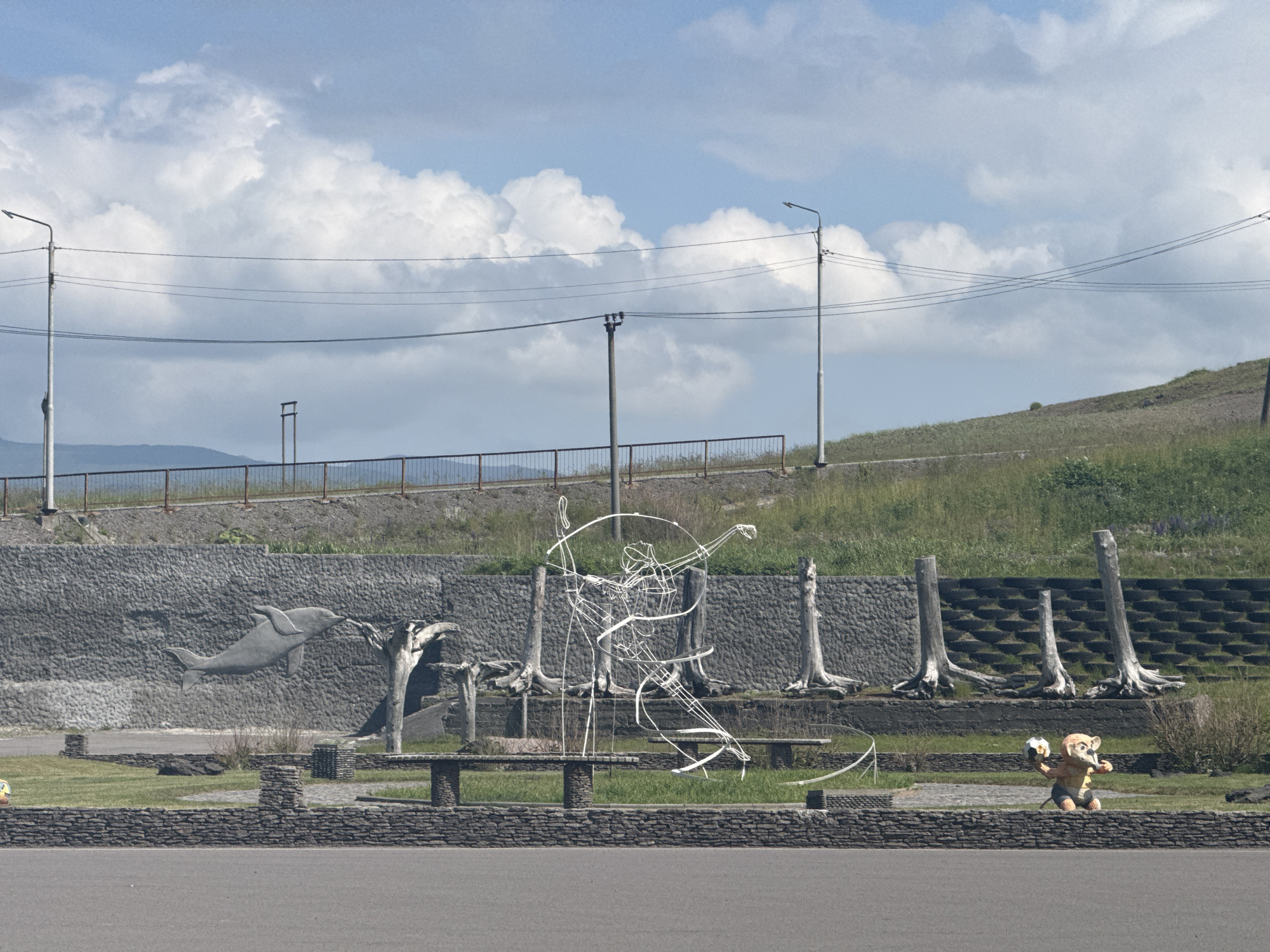 The image shows a street-level view of a park or public space in Kuril'sk, Russia, under a partly cloudy sky. In the foreground, there is a paved area, likely a road or sidewalk. Behind this, a low stone wall delineates the park area, which is covered in green grass and features several stone-built benches.
Dominating the midground is a textured stone wall with a carved relief of a dolphin leaping. To the right of the dolphin relief, several bare tree stumps are planted upright, resembling a stylized grove. In front of these stumps, a wireframe sculpture depicts a human figure in a dynamic pose, possibly a dancer or athlete. Further to the right, a small, cartoonish statue of an animal, possibly a raccoon or bear, is holding a black and white sphere, resembling a soccer ball.
The background includes a grassy embankment, bordered by a metal fence and a road. Several utility poles with power lines are visible against the sky, along with distant hills or mountains. The overall scene suggests an outdoor public space with artistic installations and natural elements.