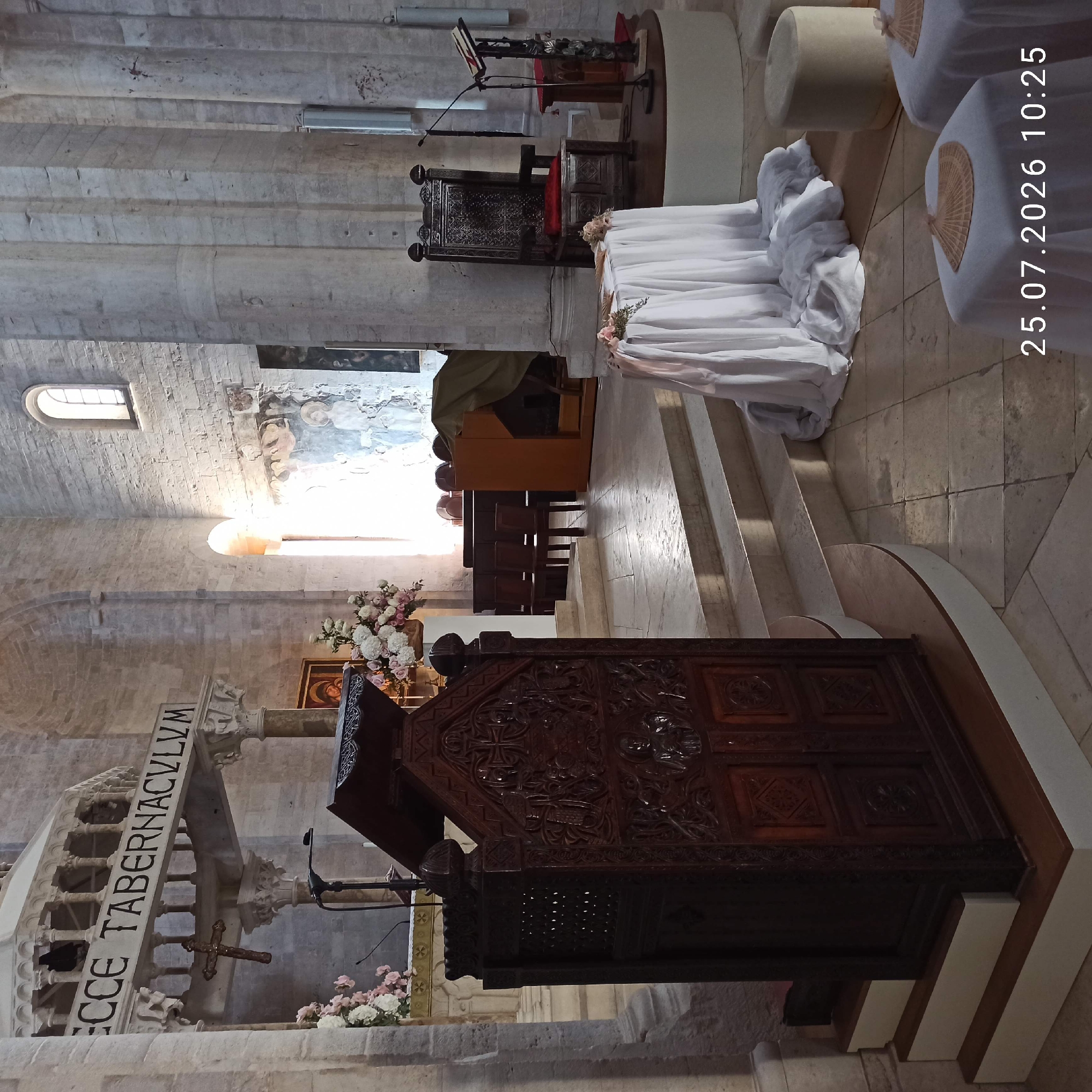 The image captures the interior of the Cathedral of Ruvo di Puglia, Italy. In the foreground, a large, ornate wooden pulpit stands on a raised platform, intricately carved with religious motifs. To the left of the pulpit, a cluster of white and pink flowers is arranged. Behind the pulpit, steps lead up to a raised altar area where a white draped table is set with flowers and decorative fans. A dark, carved wooden chair is positioned to the right of the altar. The architecture features large stone columns and arched doorways, characteristic of Romanesque style. A fresco is visible on a distant wall. The overall scene depicts the solemn and decorative ambiance of a church interior, possibly set up for a religious ceremony. The date stamp "25.07.2026 10:25" is superimposed on the lower right corner of the image.