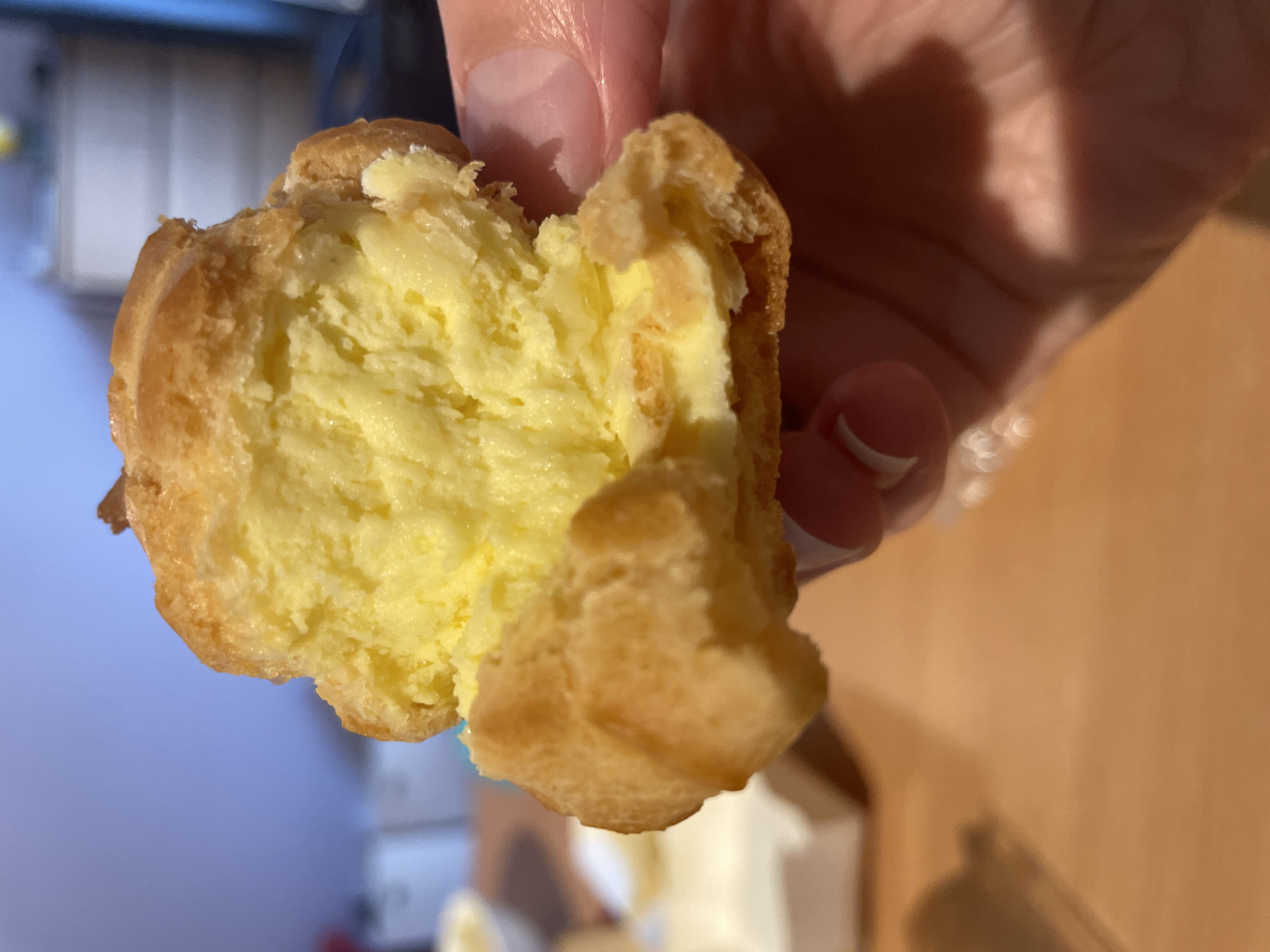 A person is holding a partially eaten cream puff. The cream puff has a golden-brown shell and is filled with a light yellow cream filling. The background is blurred and shows a room with neutral-colored walls and furniture. The location is unspecified.
