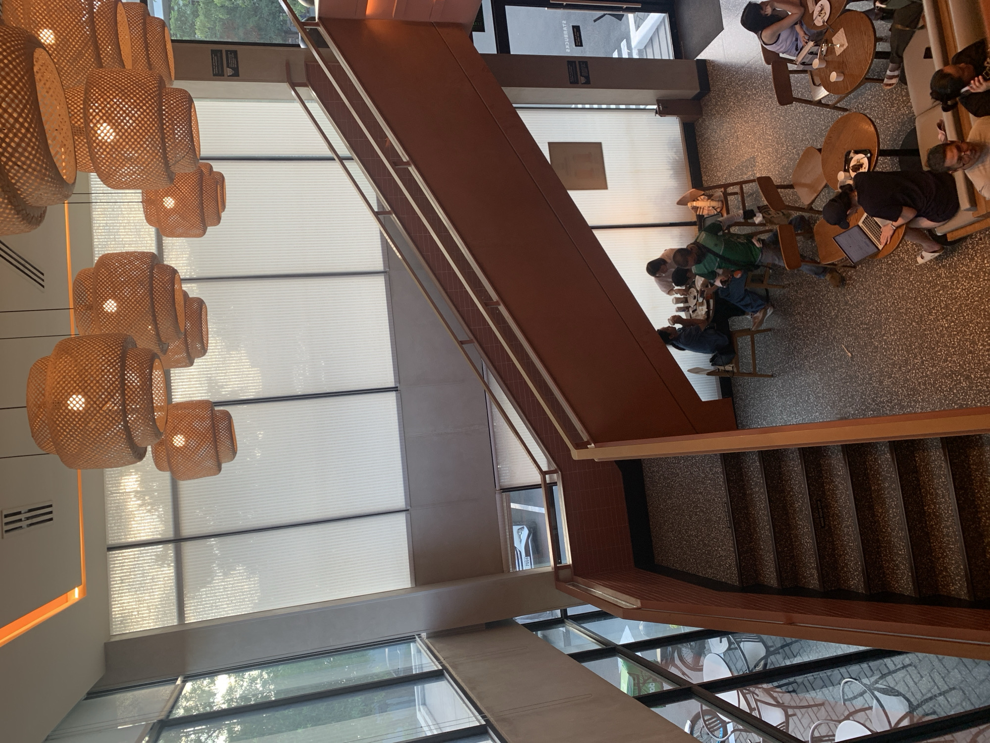 This is an indoor shot of a multi-level establishment, possibly a cafe or restaurant, in Angeles City, Philippines. The scene is captured from an elevated perspective, looking down on a staircase and an open area with seating.
Dominating the upper part of the image are several large, woven pendant lights, casting a warm glow. A grand staircase, with terracotta-colored risers and a wooden handrail, spans diagonally across the frame, leading to an upper level. The staircase has a speckled grey tread.
Below and to the right of the staircase, tables and chairs are arranged. Several people are seated at these tables, engaged in various activities. One person is working on a laptop, while others appear to be conversing and eating. A few individuals are standing or in motion, suggesting a dynamic environment.
The overall ambiance is modern and inviting, with large windows allowing natural light to filter in, creating a bright and airy atmosphere. The flooring is a speckled terrazzo, and the walls are a muted tone. To the right, a doorway with a glass panel is visible, possibly an entrance or exit.