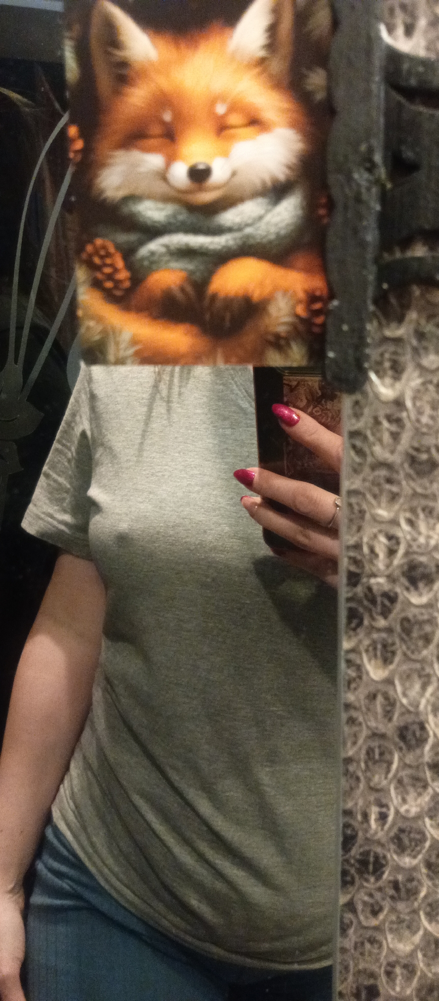 The image is a mirror selfie where a person's face is intentionally obscured by a picture of an animated fox. The fox, depicted with vibrant orange fur and a white underside, has its eyes closed in a serene, smiling expression. It is adorned with a grey-green knitted scarf and appears to be holding pinecones, conveying a cozy and charming aesthetic.
The visible portion of the person shows them wearing a plain grey short-sleeved t-shirt and blue jeans. Their right hand, adorned with long, sparkly pink-red nail polish and a subtle ring on the ring finger, holds a dark, rectangular object, likely a smartphone, used to capture the reflection.
The setting appears to be indoors, indicated by the mirror and the surrounding elements. On the left side of the reflection, a dark and blurry background is visible. To the right, a textured surface, resembling bubble wrap or a patterned wall, is reflected. The lighting suggests an indoor environment, with no discernible time of day or specific location cues beyond the general interior. No text is visible in the image.