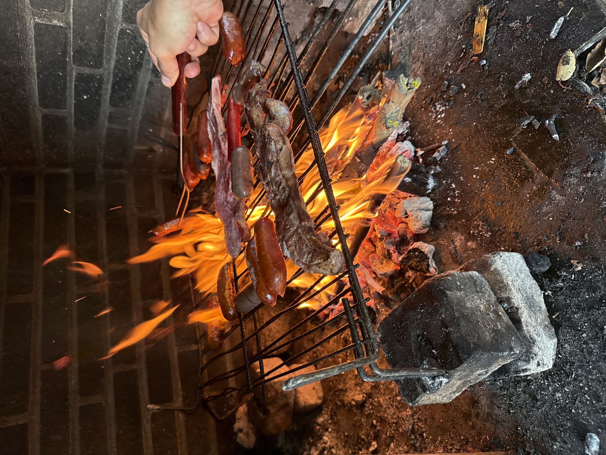 A close-up, eye-level, outdoor shot shows a barbecue grill with various meats cooking over a vibrant fire in Santa Cristina d'Aro, Spain. The grill grates are laden with several sausages and pieces of steak. A hand holding a grilling tool is visible on the right side, actively tending to the food. The background features a rustic brick wall, with visible flames and glowing embers beneath the grill, suggesting a lively cooking process. The ground around the grill is covered in ash and debris, indicating recent or ongoing use.