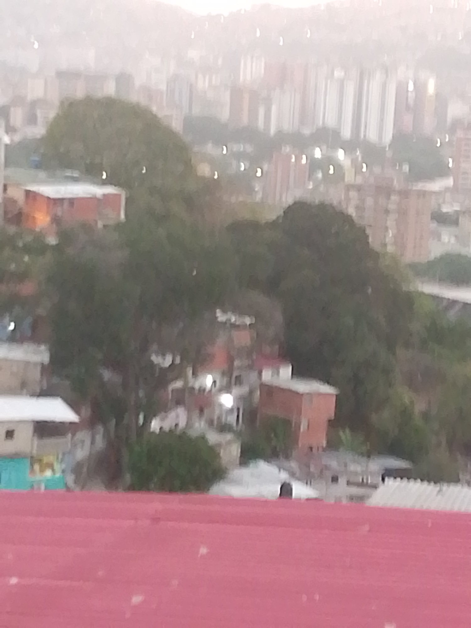 The image shows a blurry urban landscape in Caracas, Venezuela, characterized by hills covered with trees and densely packed residential buildings. In the foreground, a red roof dominates the lower portion of the image. The background reveals numerous tall apartment buildings, suggesting a high-density urban area. The overall atmosphere appears hazy, possibly due to smog, rain, or fog. The scene lacks identifiable people or specific actions.