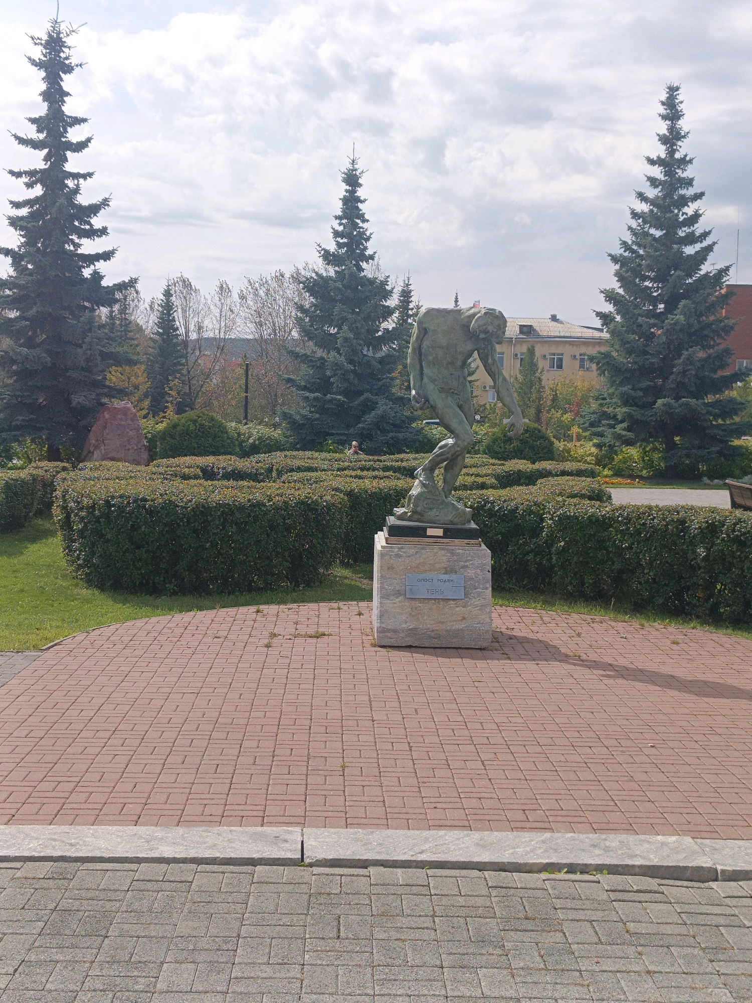 The image depicts an outdoor scene in the city of Satka, Russia, featuring a bronze sculpture on a stone pedestal. The sculpture, which appears to be "The Thinker" by Auguste Rodin, is positioned on a circular, reddish-brown brick paved area. Behind the sculpture, a meticulously maintained hedge forms a maze-like garden with a large, reddish-brown boulder. Several tall, dark green coniferous trees frame the scene, with a light-colored building and a red-brick building visible in the background beyond the trees. The sky is partly cloudy with a soft, diffused light. The ground in the foreground is paved with grey bricks. The overall impression is of a public park or square.