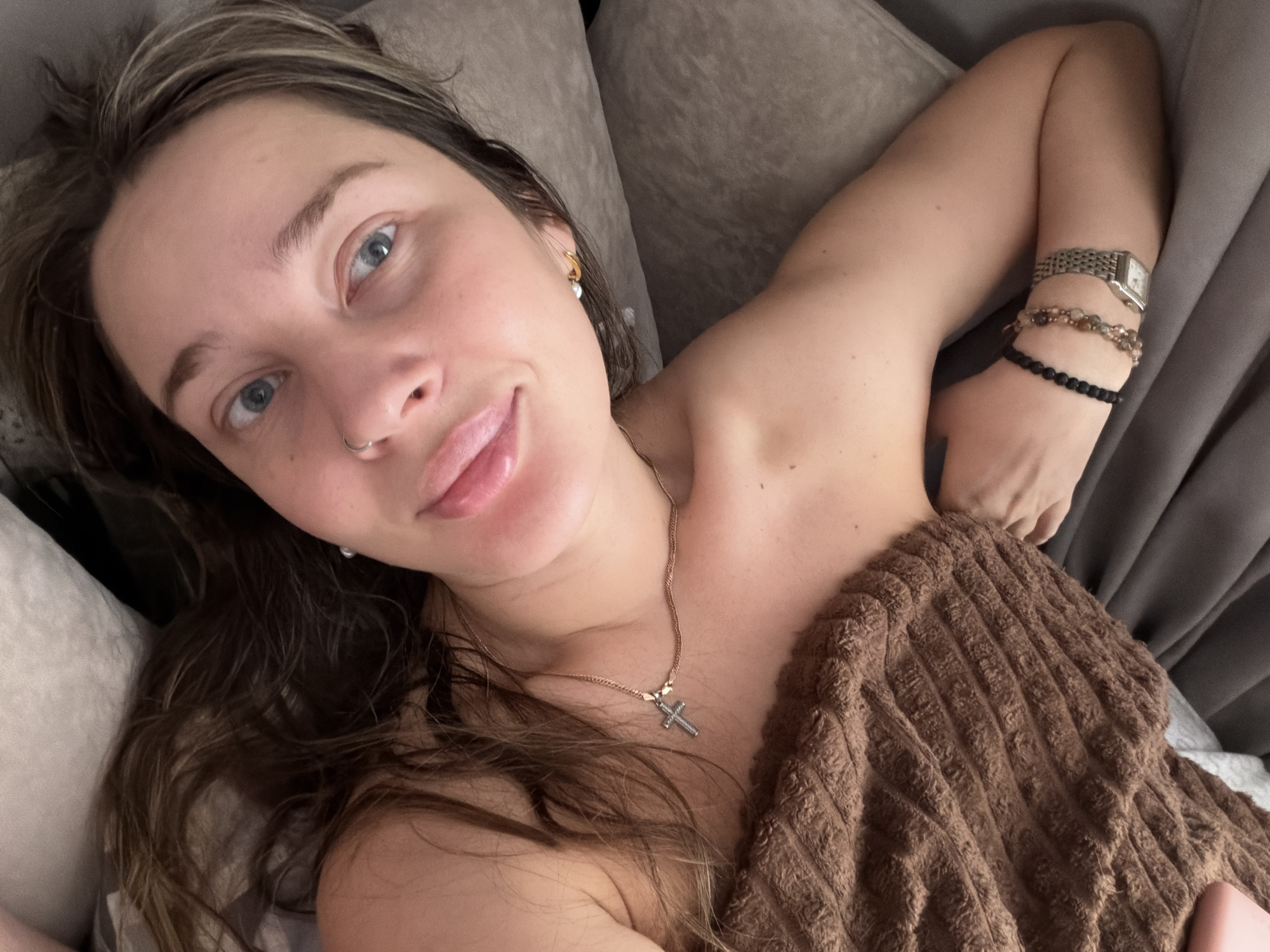 A woman is lying on a grey upholstered surface, possibly a couch or bed, and is partially covered by a brown ribbed blanket or throw. She has light skin, light brown hair with blonde highlights, and blue eyes. She is wearing a nose ring, stud earrings, and a necklace with a silver cross pendant. On her wrist, she has a silver watch and two beaded bracelets. The location is not discernible from the image.