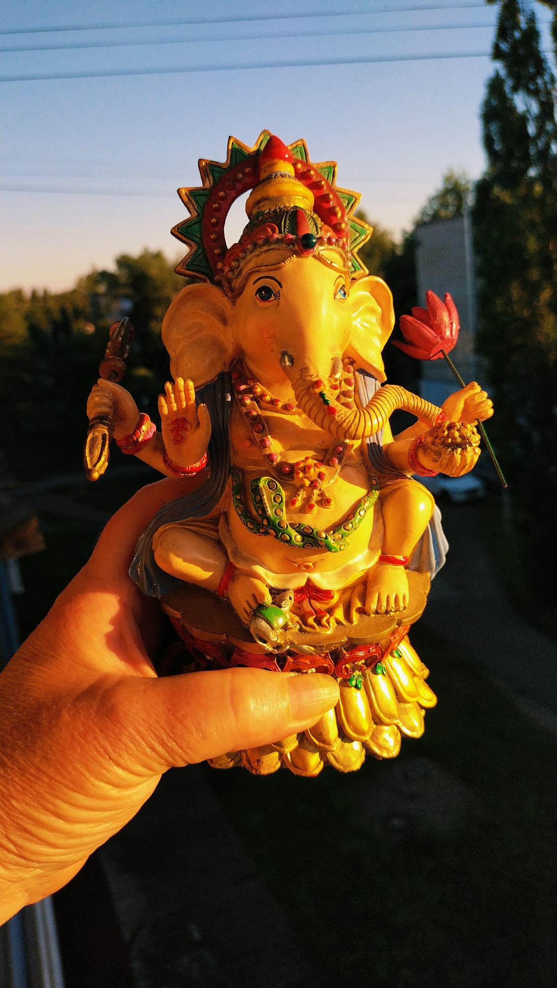 An overhead shot captures a brightly colored statue of Ganesha being held by a person's hand. The statue is yellow and adorned with red, green, and gold details, including jewelry and a headdress. Ganesha holds a lotus flower in one hand. The background is a blurred image of trees and a building, with a clear blue sky above. The lighting suggests it is either early morning or late afternoon, with the sun casting a warm glow on the statue and hand. The scene appears to be outdoors, possibly on a balcony or near a window, given the presence of what looks like a railing in the bottom left corner. The specific location in Horishni Plavni, Ukraine, is not visually indicated in the image itself.