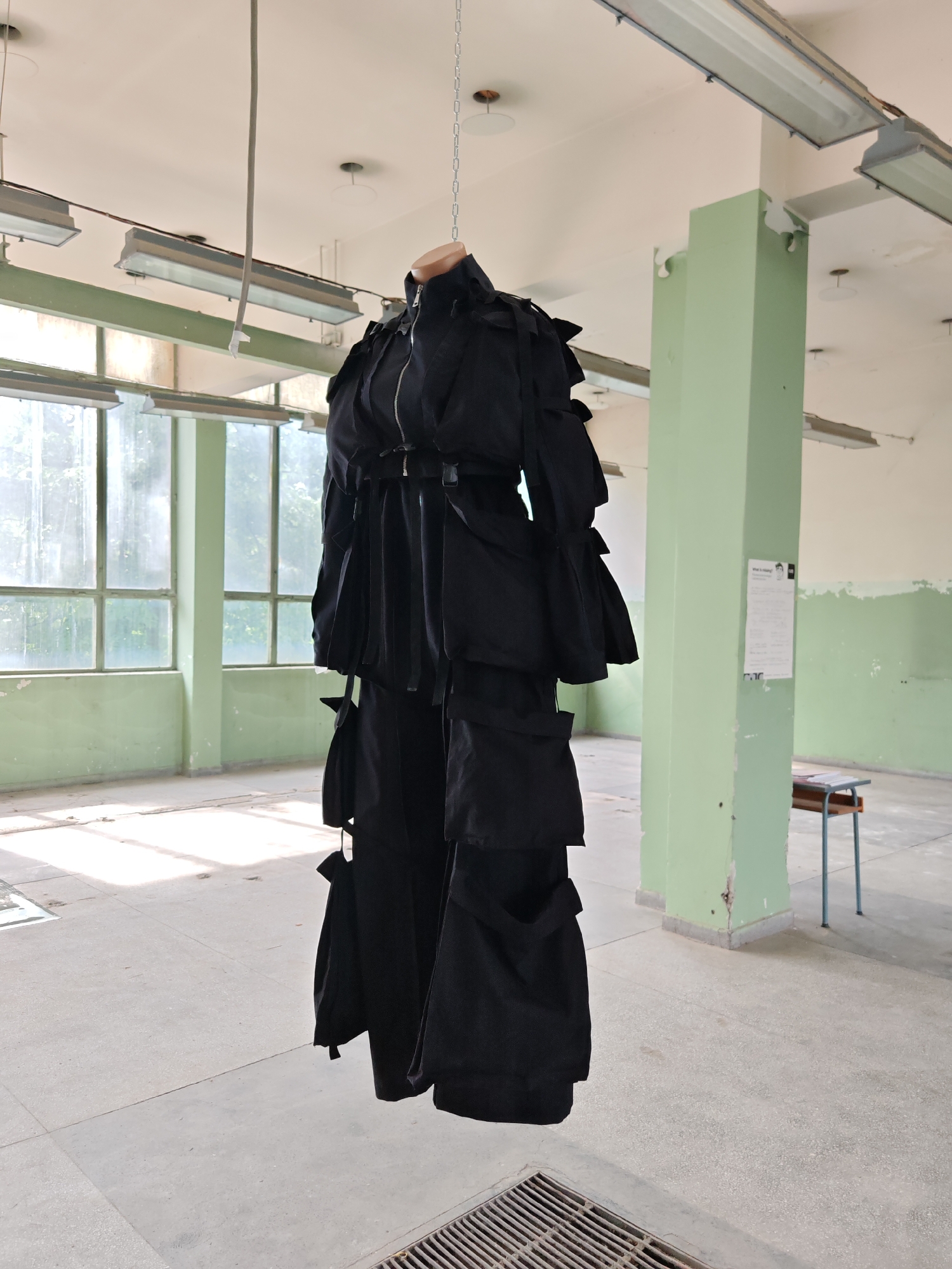 The image captures an interior scene within a large, disused or semi-abandoned building in Gabrovo, Bulgaria. A dark-colored, multi-pocketed garment, resembling a coat or jumpsuit with a zippered front and numerous straps and buckles, is suspended centrally from the ceiling by a silver-colored metal chain. The garment's upper section includes a light-colored, mannequin-like neck form.
The room exhibits characteristics of an aged industrial or institutional structure. The ceiling is off-white and features multiple exposed fluorescent tube light fixtures mounted in grey metal housings. Several circular, recessed ceiling elements are also present. A single grey cable hangs detached from the ceiling on the upper left. The walls are demarcated into two distinct paint sections: an upper cream or off-white and a lower pale mint green, prominent on a large structural column located in the mid-ground right. This column displays chipped paint and surface irregularities. The left wall is dominated by large, multi-paned windows with light-colored frames, admitting substantial natural light. Exterior foliage is visible through the windows.
The floor is a light grey concrete or large-tiled surface, showing signs of wear and dirt, with areas of strong direct sunlight and shadows. A metal floor grate is visible in the bottom right foreground. In the background, to the right of the mint green column, a small, aged wooden desk with blue-grey metal legs is present. A white poster, featuring indistinct text and an image of a person, is affixed to the right side of the mint green column. No individuals are present in the scene. The garment is static, suspended vertically.