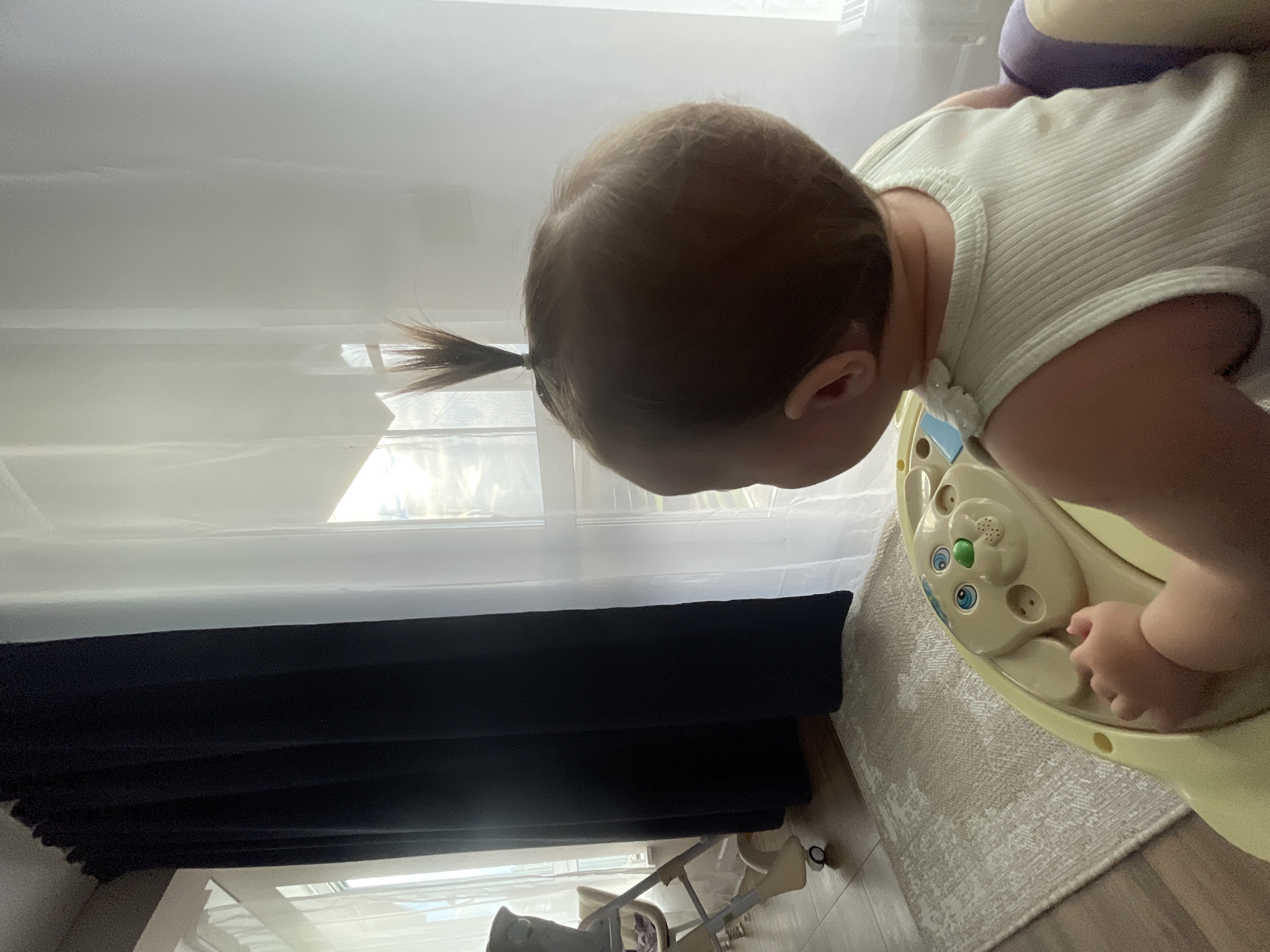 The media file shows a close-up, low-angle shot of a toddler from the side, facing right. The toddler has light brown hair pulled into a small ponytail. They are wearing a light green ribbed tank top and appear to be leaning over a yellow toy. The toy is shaped like a bear's face with blue eyes, a green nose, and various buttons and features. The toddler's right hand is visible, reaching towards the toy.
The background features a sheer white curtain, suggesting the presence of a window, likely letting in natural light. Below the curtain, a dark black fabric is visible, possibly a drape or shade. The floor appears to be light-colored wood with a textured rug on top. In the lower part of the frame, a white baby rocker or similar baby equipment is partially visible.
The overall scene suggests an indoor setting, possibly a living room or playroom, during the daytime, indicated by the natural light. The image appears to be taken from a floor-level perspective, emphasizing the toddler's interaction with the toy. There is no visible text in the media file. The focus is on the toddler and the toy, creating an intimate and possibly playful mood. The image is oriented sideways.