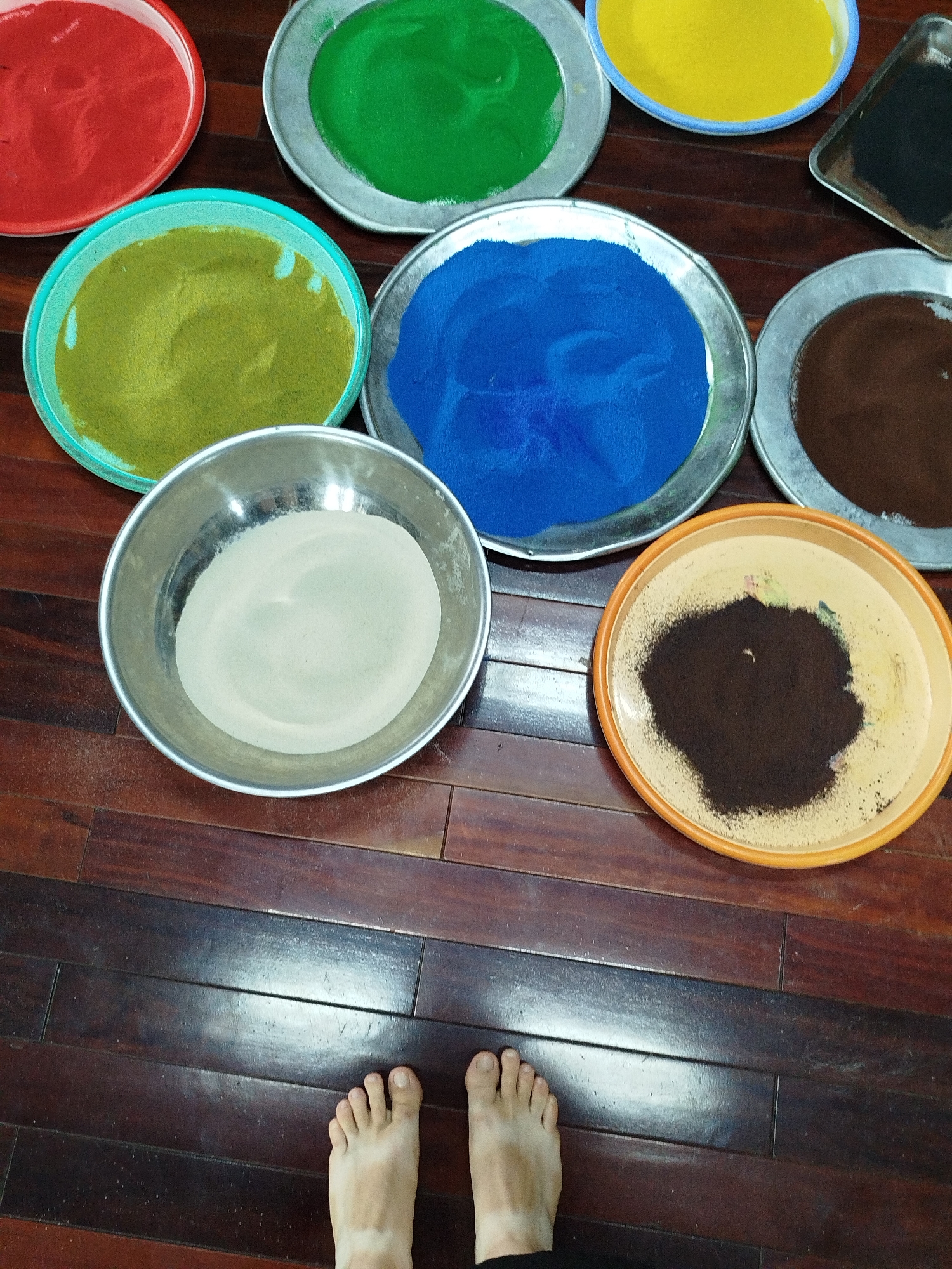 An overhead shot shows a collection of eight shallow dishes, each filled with a different colored sand, arranged on a polished dark wood floor. The dishes are in various circular and square shapes, and the sand colors include red, green, yellow, blue, olive green, light gray, dark brown, and a very dark brown. The red, green, yellow, and blue sand dishes appear to be larger and contain a significant amount of sand. The dish with light gray sand is a metal bowl and appears to be filled with a fine powder. The two brown sand dishes are smaller and contain less sand. In the lower portion of the image, two bare feet with light skin are visible, placed on the wooden floor. One foot is turned slightly towards the right, and the other is mostly obscured by the bottom edge of the frame. The lighting in the scene is bright, casting reflections on the polished floor. The setting does not provide specific visual cues to pinpoint a location within Da Lat, Vietnam.