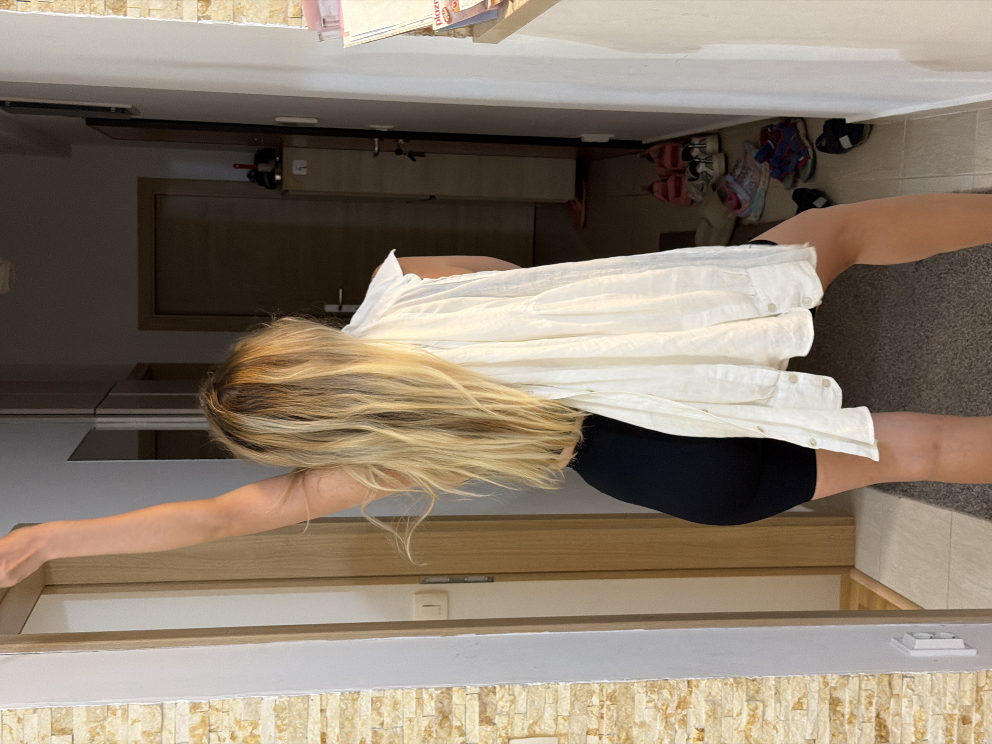 A woman with blonde hair is standing in a hallway, facing away from the camera. She is wearing black shorts and a white shirt draped over her shoulders. Her right arm is raised, touching the top of the doorway. The hallway is decorated with light-colored stone on the left wall and white walls on the right. There are several doors visible in the background, as well as a collection of shoes on the floor near the right wall. The lighting suggests it is daytime. The scene appears to be indoors, possibly in a residential building. The location of Budva, Montenegro, is not evident from the image itself.
