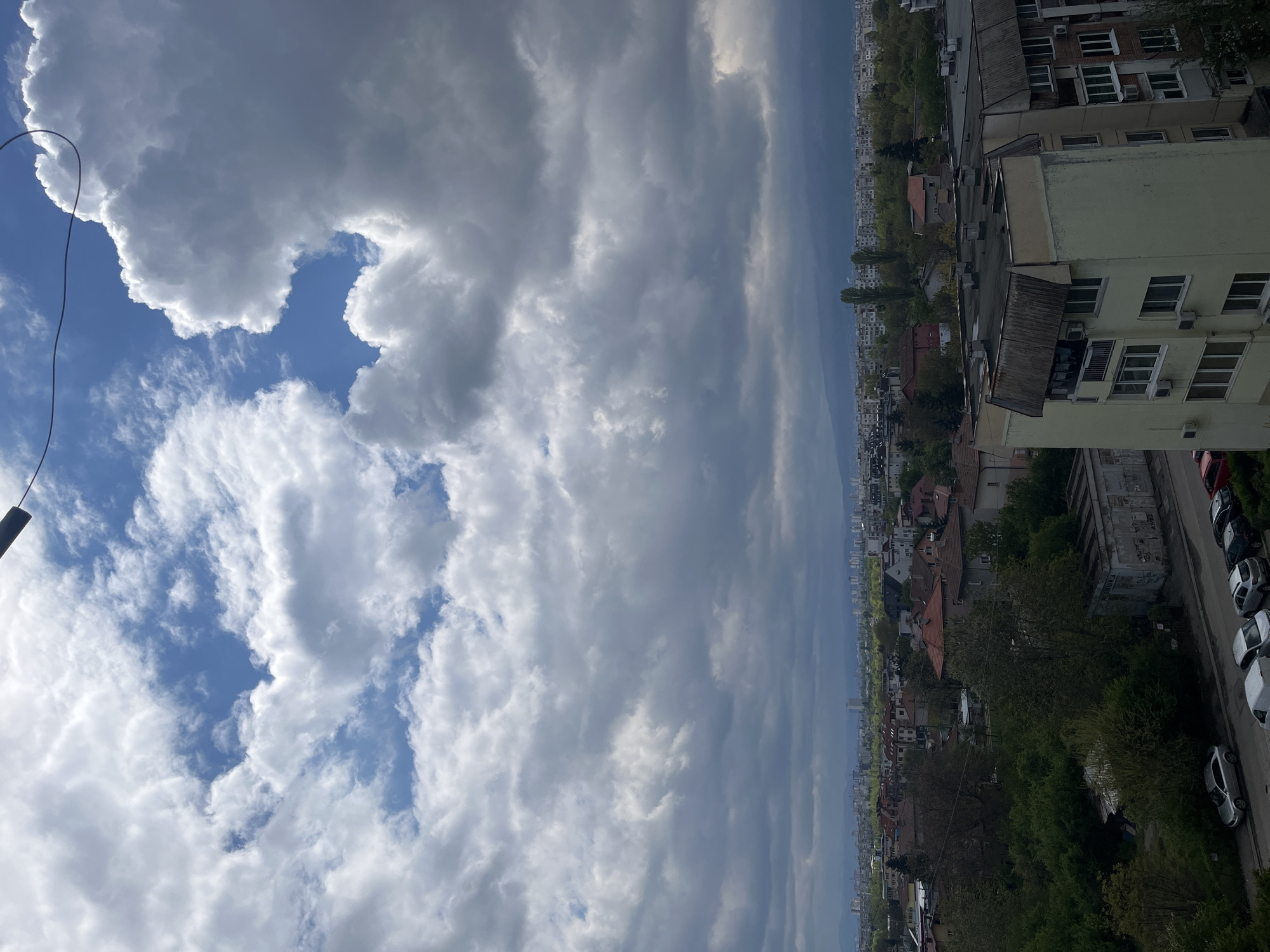 The image presents a view from a high vantage point, likely a building in Sofia, Bulgaria. The sky dominates the upper portion of the frame, showcasing a mix of cumulus clouds and patches of blue sky. Below, the urban landscape extends into the distance, featuring a mix of residential buildings with visible windows, rooftops, and some with air conditioning units. The cityscape also includes pockets of green space with trees. A street runs along the edge of the building with a line of parked cars, which appear to be mainly white and gray. A cable or wire is visible in the top left corner of the frame. Overall, the scene depicts a typical urban environment in Sofia under a partially cloudy sky.
