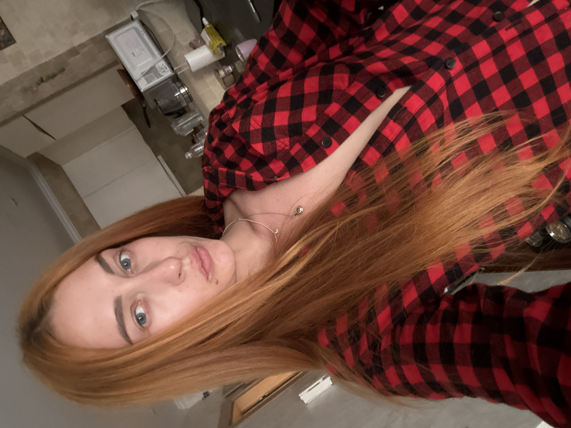 A woman with long, reddish-auburn hair and blue eyes is depicted in a close-up, vertically oriented portrait, likely a selfie. She looks directly at the camera with a neutral expression, her lips slightly pursed. She is wearing a red and black buffalo plaid button-down shirt, unbuttoned at the top, revealing a part of her chest and two delicate silver-toned necklaces, one with a small, circular pendant. Her hair cascades around her shoulders and over parts of her shirt.
The background suggests an indoor setting, possibly a kitchen or utility area in Budva, Montenegro. To the woman's upper right, a light-colored countertop is visible, holding various indistinct items including bottles, a white rectangular appliance, and another kitchen device. The surrounding walls or cabinets appear in muted, light tones. There is no visible text in the image.