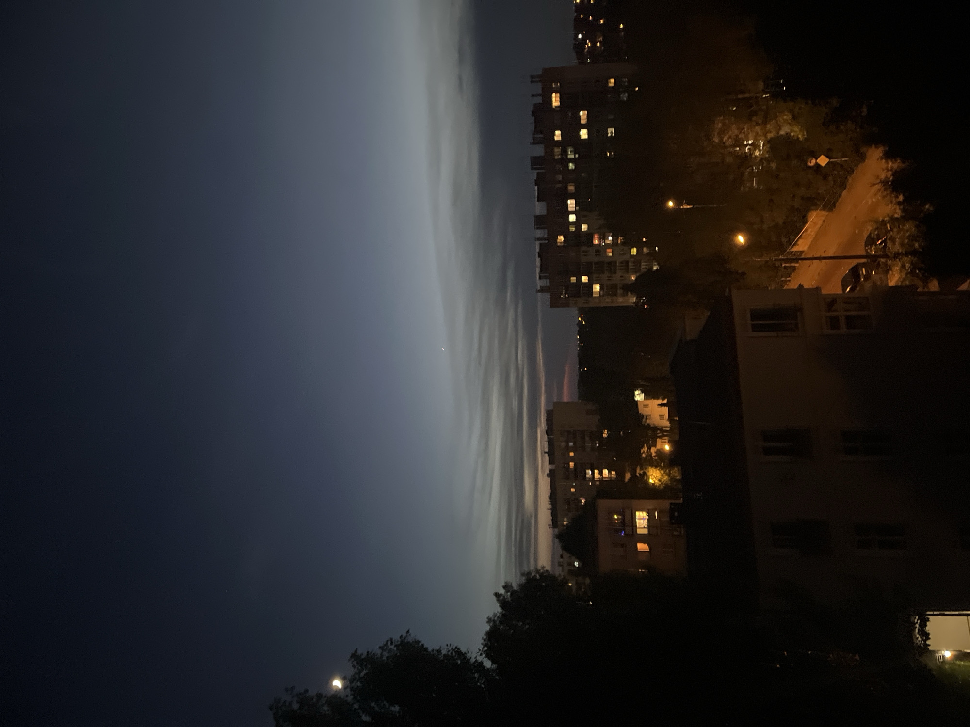A nighttime aerial view captures an urban landscape with apartment buildings and trees under a dusky sky. A sliver of a crescent moon is visible in the upper left quadrant of the sky. The sky transitions from a deep blue at the top to lighter shades of gray and faint pink near the horizon, with wispy clouds stretched horizontally across the frame. A faint pinpoint of light, possibly a distant star or planet, is noticeable in the upper portion of the sky.
Dominating the mid-ground is a multi-story apartment building with numerous windows illuminated by warm yellow light, suggesting interior occupancy. To the left and in the foreground, another building is partially visible, also with lit windows. Trees, silhouetted by the dark sky, are interspersed throughout the scene, with some appearing denser and darker than others.
A street, illuminated by streetlights, runs through the scene, with a car parked on its side. A yellow diamond-shaped road sign is visible on the right side of the street. The overall impression is of a quiet urban evening as night settles in.