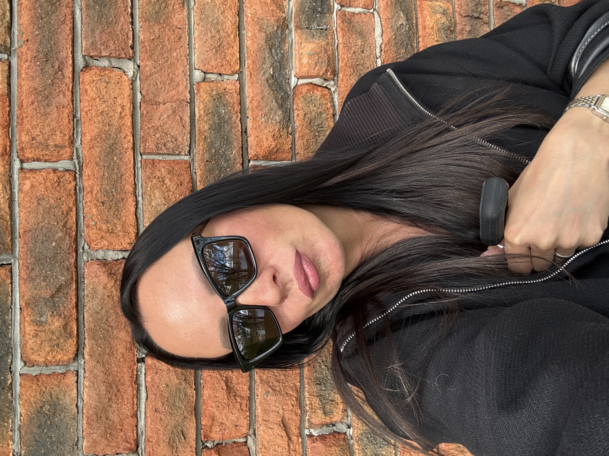 A woman with long dark hair wearing sunglasses and a black zippered jacket is posing for a picture in front of a brick wall. She has a watch on her wrist. The location of the photo is not discernible from the provided image. It is not possible to verify that this image was taken in the city of Chaikovsky, Russia.