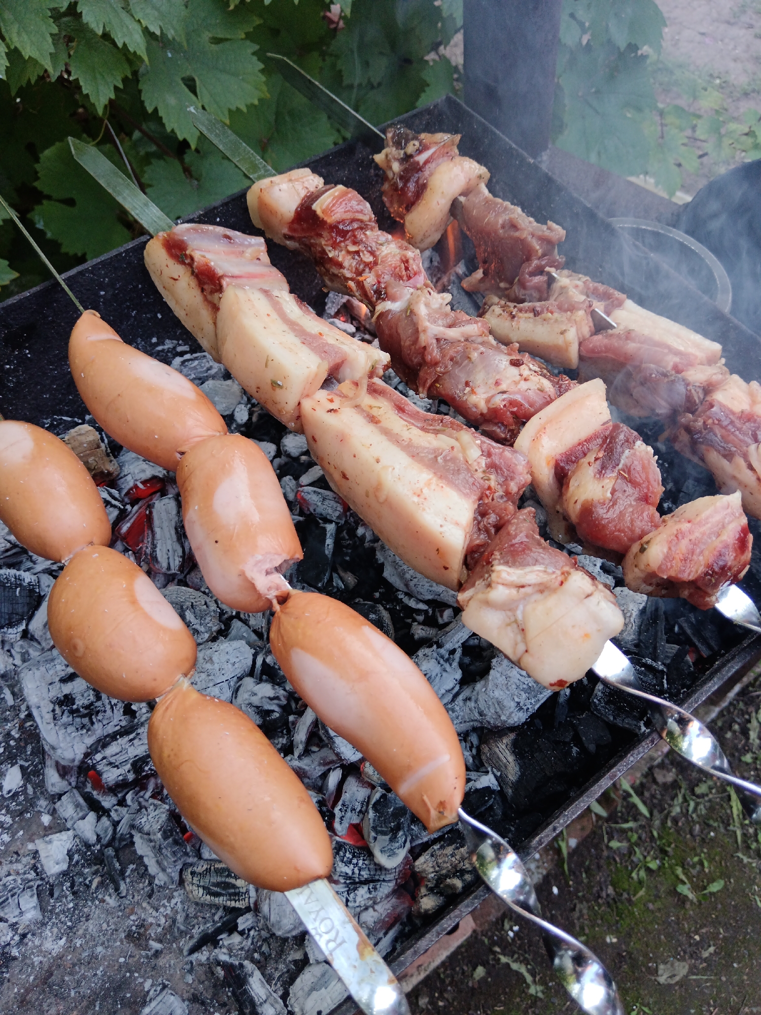 The image depicts an outdoor barbecue scene, likely in Sorochinsk, Russia, given the context. In the foreground, several skewers of sausages are arranged on a charcoal grill. The sausages are a uniform light brown color and appear to be fully cooked or nearing completion. Behind the sausages, skewers of raw meat, predominantly pork with visible layers of fat and lean muscle, are cooking over glowing embers. Wisps of smoke rise from the grill, indicating active cooking. The grill itself is a dark metal, rectangular structure filled with charcoal and ashes. In the background, green foliage, possibly from a garden or trees, provides a natural backdrop. The ground surrounding the grill is covered with dirt and grass. The scene evokes a casual outdoor dining or gathering atmosphere.