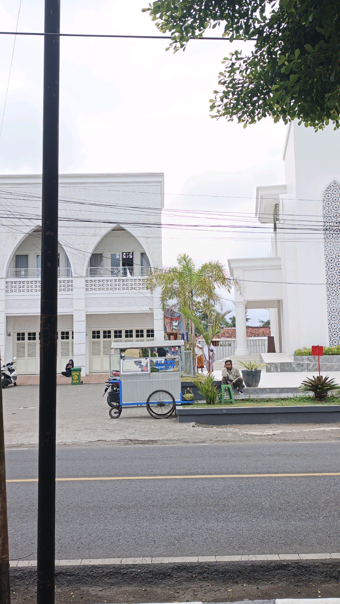 A street scene in Desa Karangbenda, Indonesia, features a modern white building with arched windows and decorative white latticework. In front of the building, a man sits on a low wall, looking to his right. To his left, a small food cart with a blue and silver body and bicycle wheels is parked. A person in a green hijab is seated near the cart. A white scooter is parked further to the left. The street in the foreground is asphalt with a yellow dividing line. Overhead, green leaves from a tree hang into the frame.