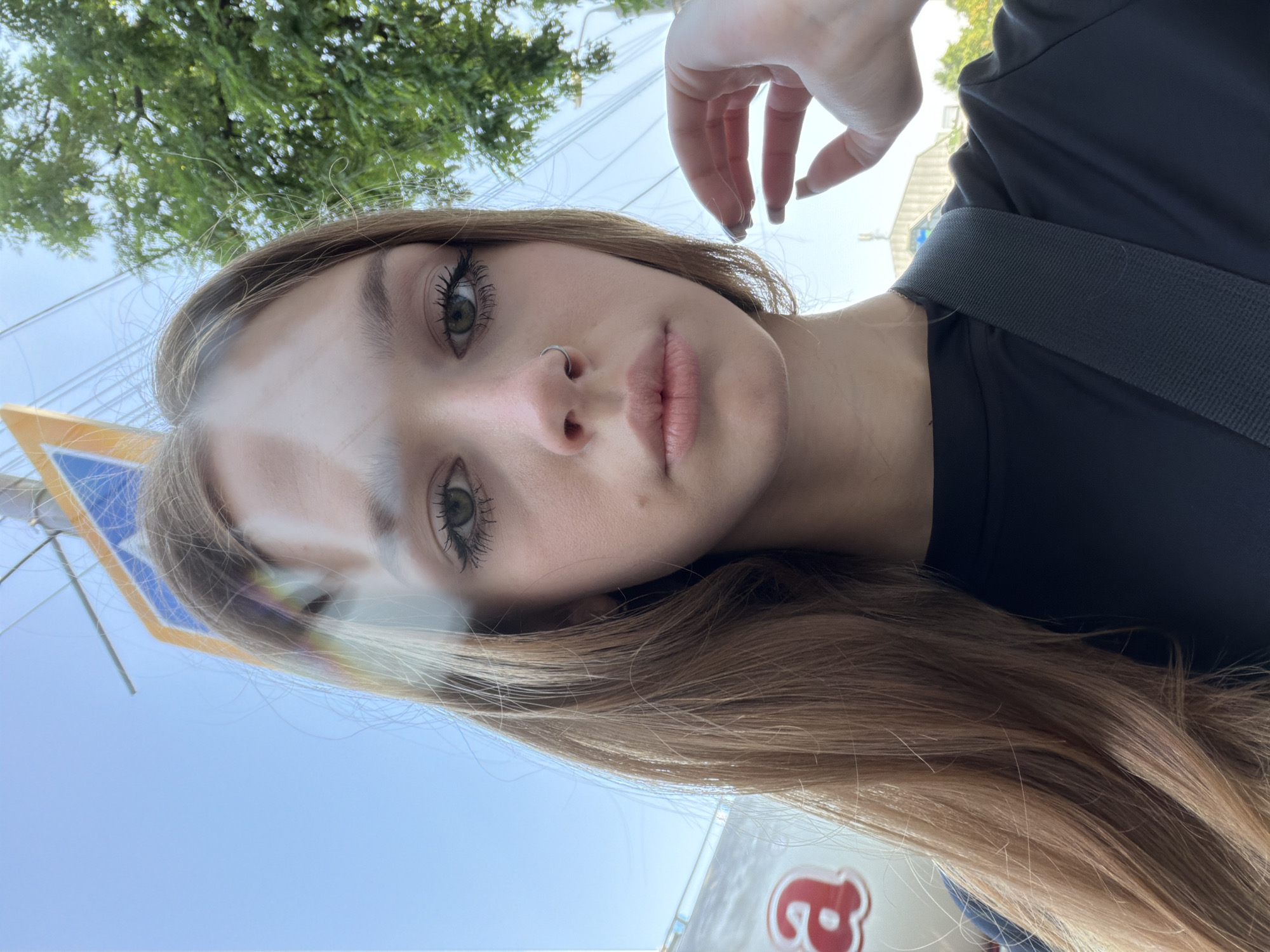 A young Caucasian woman with long, light brown hair is captured in a close-up selfie taken outdoors. She is wearing a black top and has a silver hoop nose ring and a nose piercing. Her right hand is raised, with her fingers near her face, and she is wearing dark nail polish. The background includes a street sign, a tree with green leaves, power lines, and a blurry vehicle with a red logo on its side. The sky is blue.