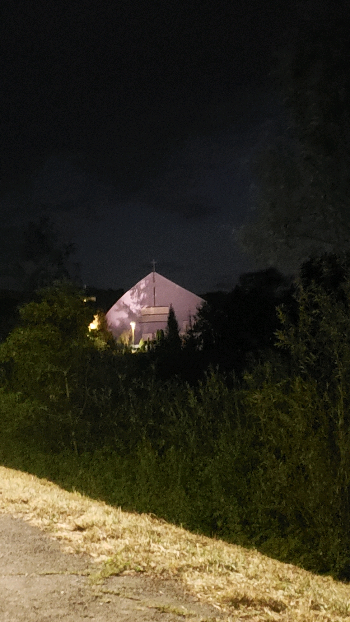 An evening photograph shows a modern church building with a triangular roof and a cross at its apex, situated in the city of Prešov, Slovakia. The church is illuminated with a pinkish light, casting shadows on its facade. It is surrounded by dark trees and foliage, with a small amount of yellowed grass and a paved surface visible in the foreground. The sky above is dark and cloudy, suggesting nighttime or twilight. There are no people visible in the image.