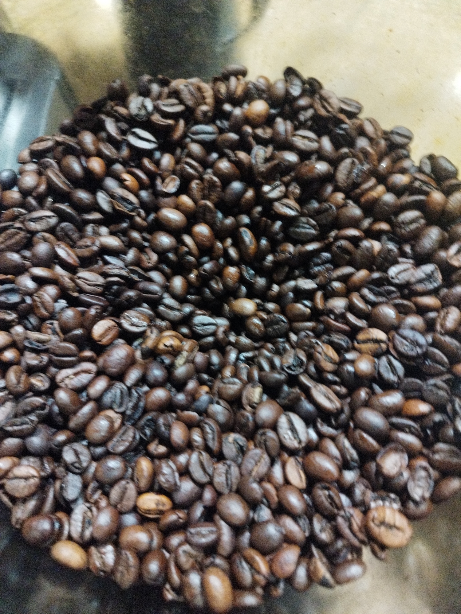 This is a close-up, overhead shot of a large pile of roasted coffee beans in what appears to be a transparent container, possibly a coffee grinder hopper or a storage bin. The coffee beans are dark brown and vary in shade, with some showing a lighter brown hue and a visible crack down the middle. They are densely packed and create a textured surface with shadows and highlights indicating their shape and depth. The lighting suggests an indoor setting, potentially a kitchen or a café, with a slightly blurred background that hints at a metallic or glass surface. There is no visible text in the image. The focus is entirely on the coffee beans, emphasizing their abundance and roasted quality. The overall impression is one of readiness for brewing, evoking the aroma and anticipation associated with coffee.